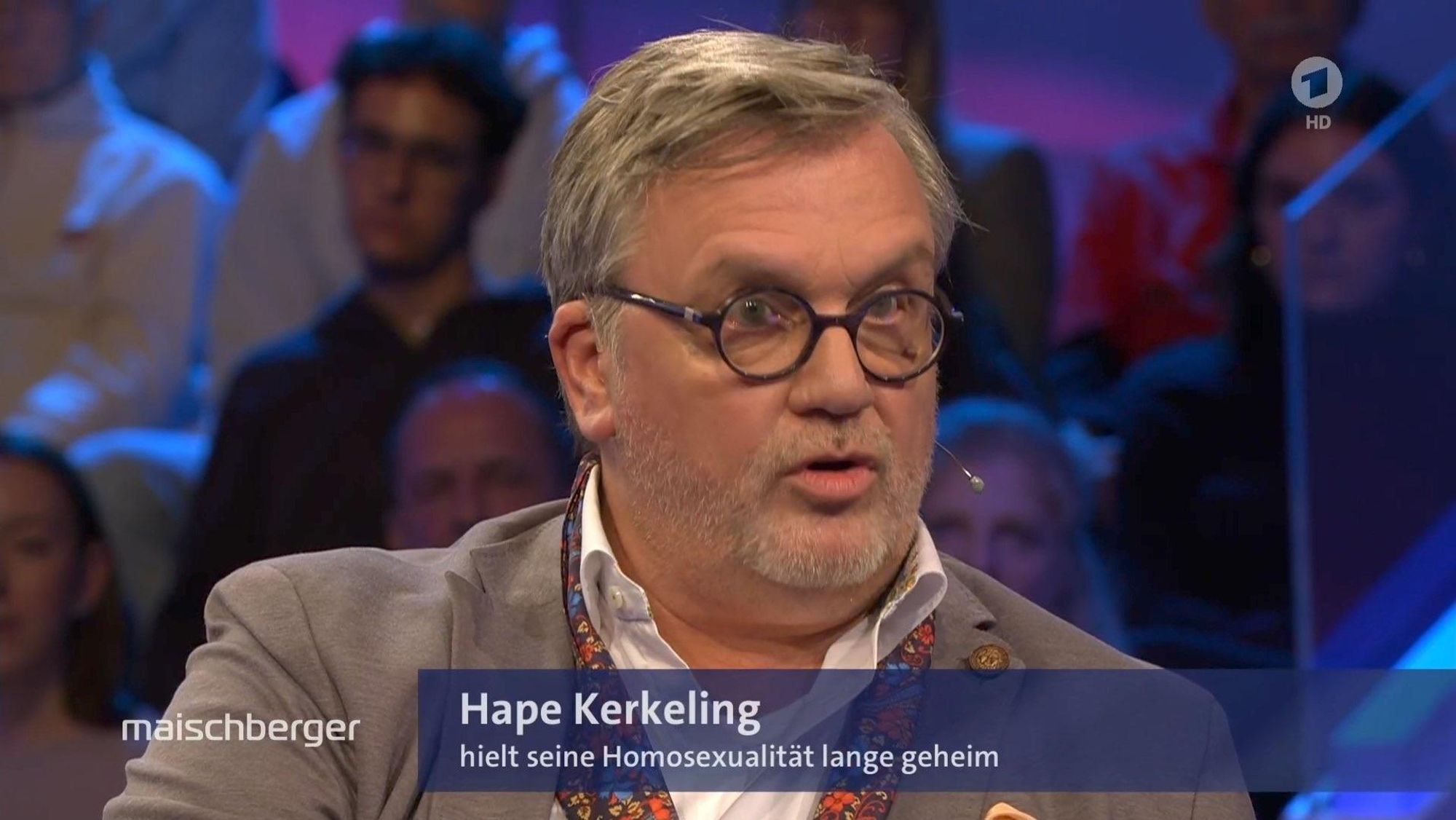 Hape Kerkeling mit grauen Haaren und Brille spricht intensiv in einem Fernsehstudio. Er trägt einen hellen Blazer mit einem dekorativen Schal. Im Hintergrund sind Zuschauer sichtbar.