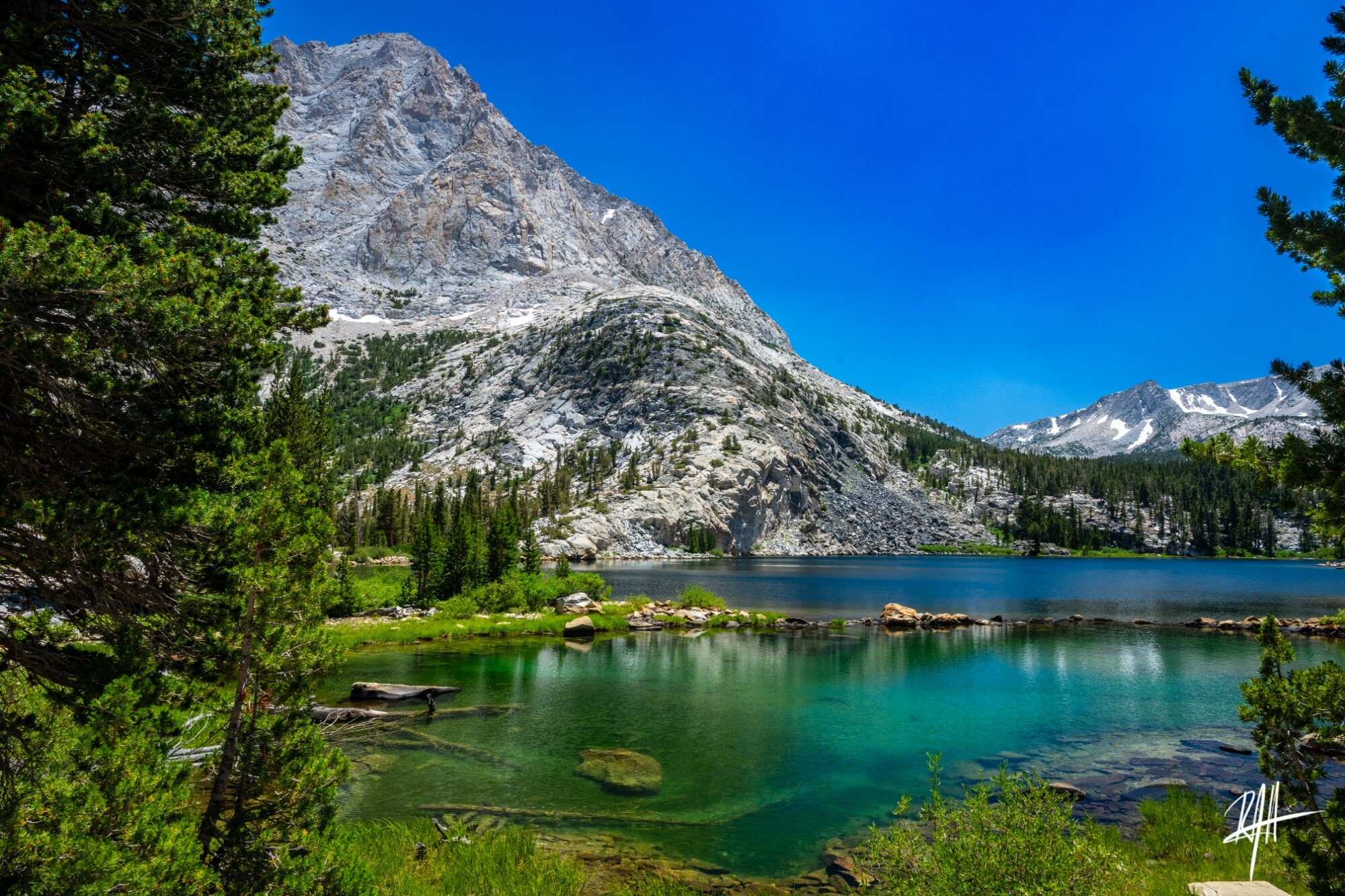 Turquoise Pine Lake framed by pine trees and steep mountains in the background with lighter felsic rocks and darker mafic rocks touching one another in the background. This is a view to the North.
