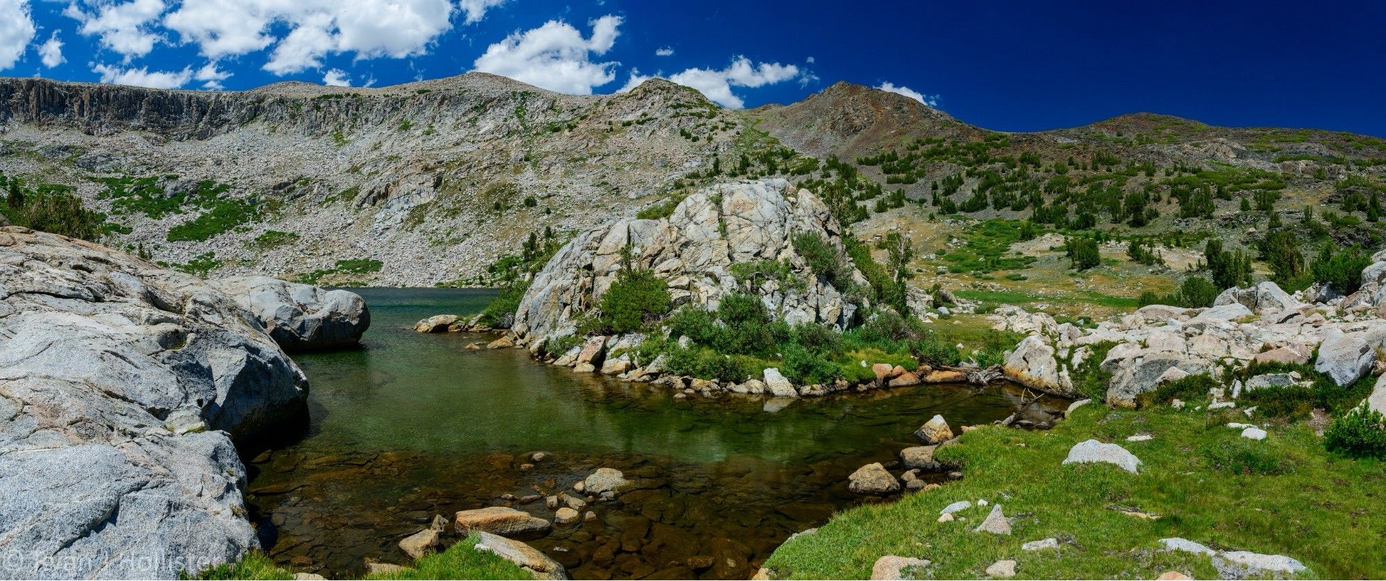 Panoramic photo of Granite lake in eastern Yosemite. Granitic landscape with a small finger shallow and clear lake in foreground with granite knob in center of photo and meadow like low grasses growing on right side of photo