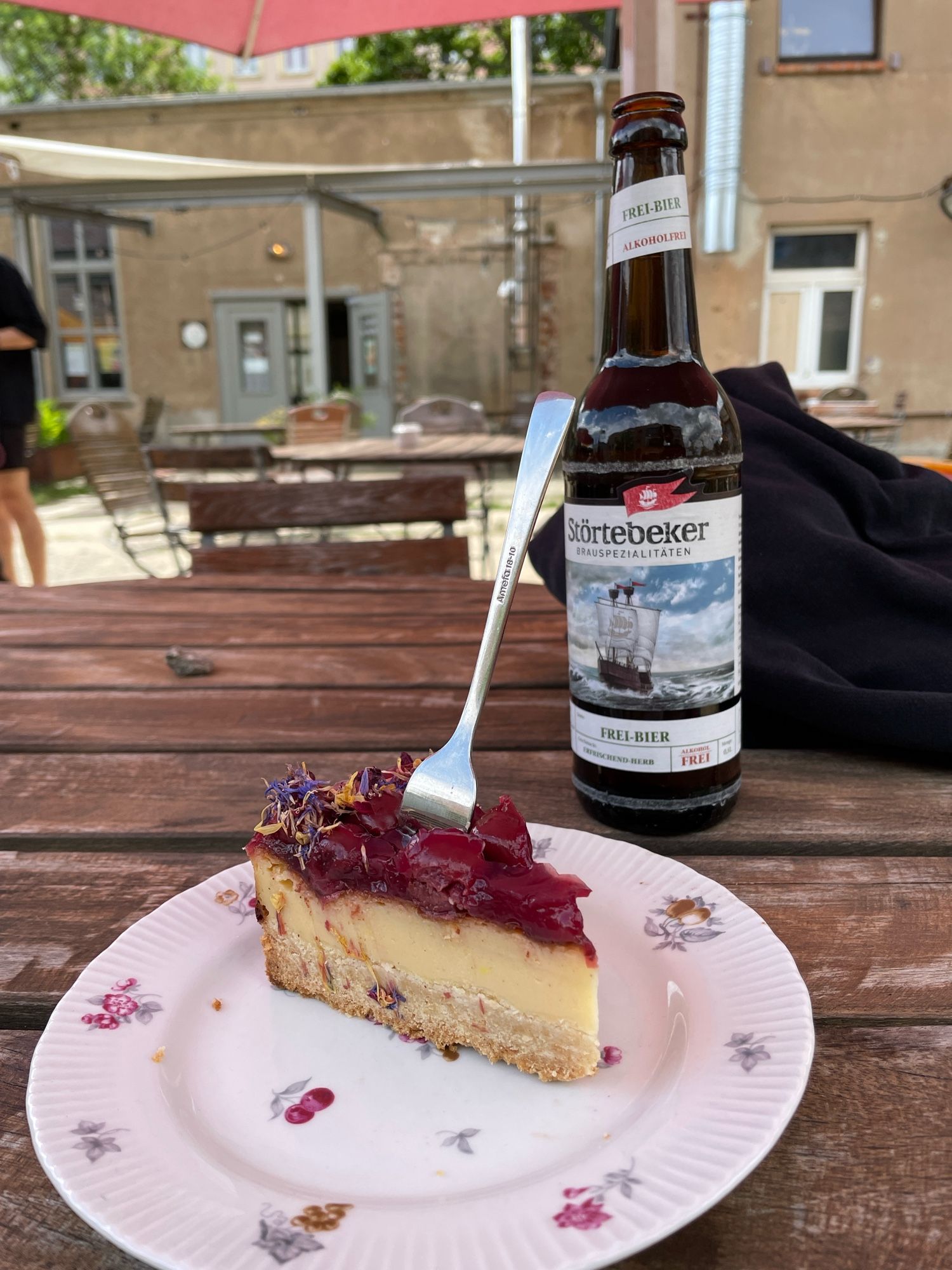 Auf einem typischen Gartentisch steht ein Teller mit Cheesecake mit Kirschen oben drauf. Dahinter eine Flasche Bier der Brauerei Störtebecker.