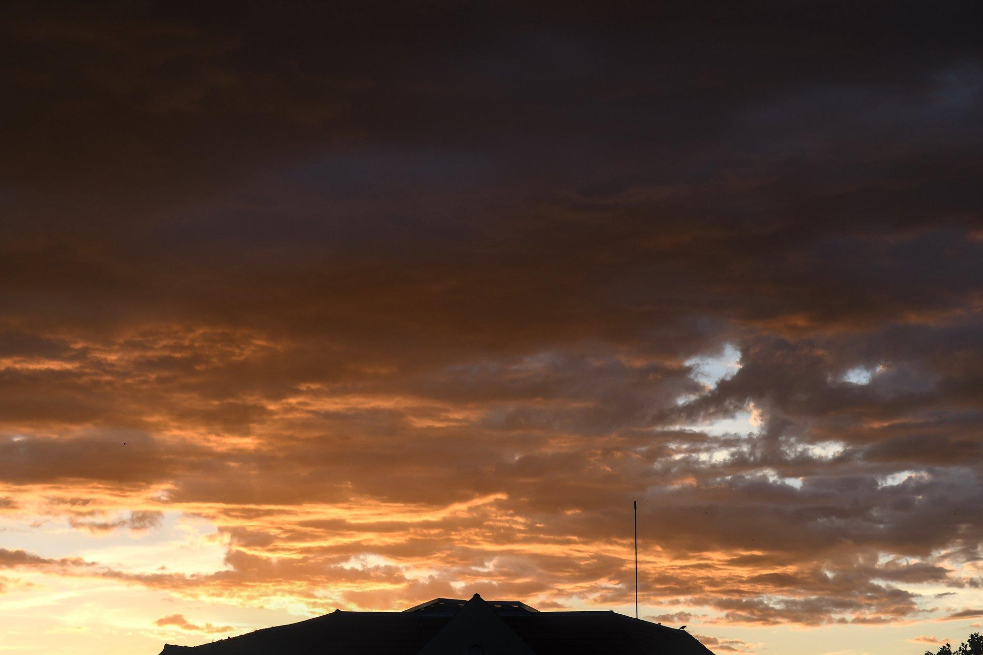 Sonnenuntergang bei stark bewölktem Himmel. Die Sonne taucht die niedrig hängenden Wolken in Gold-, Orange- und Rottöne, zum Betrachter hin erscheinen sie allerdings zunehmend bedrohlich dunkeler. Am unteren Bildrand ist das Dach des gegenüberliegenden Hauses zu sehen, auf dem ein paar Vögel sitzen.