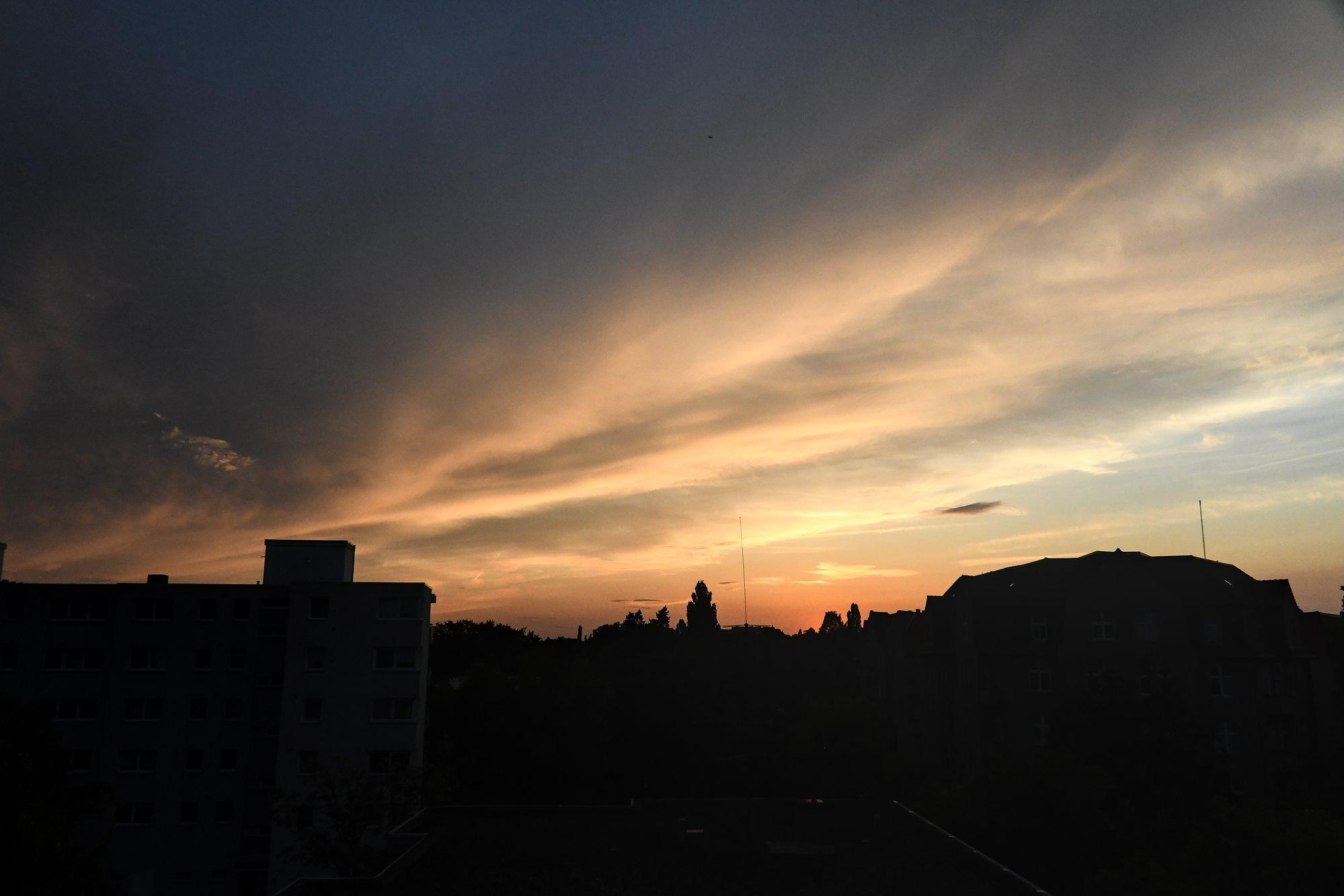 Wolkenbänder am Himmel rollen von links nach rechts. Die Sonne ist gerade untergegangen, sodass die Wolken von unten angeschienen werden. Halb als Silhouetten sind Wohnhäuser der Nachbarschaft und Bäume im Park auf der anderen Seite der Hauptstraße zu sehen.