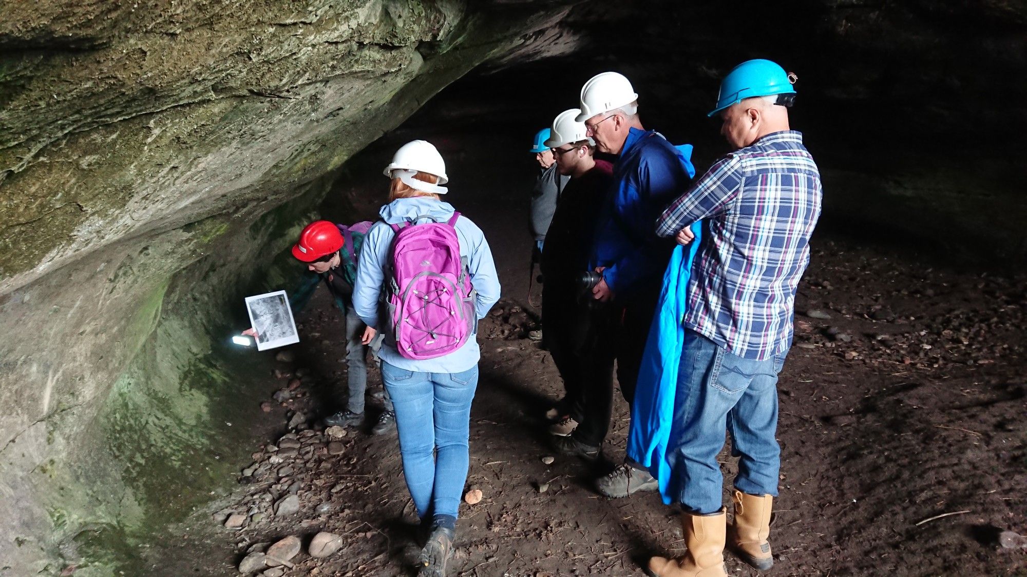 A group of people in hard hats, standing in a cave. A person in a red hard hat is bending, holding a picture and shining a light on the cave wall to the left.