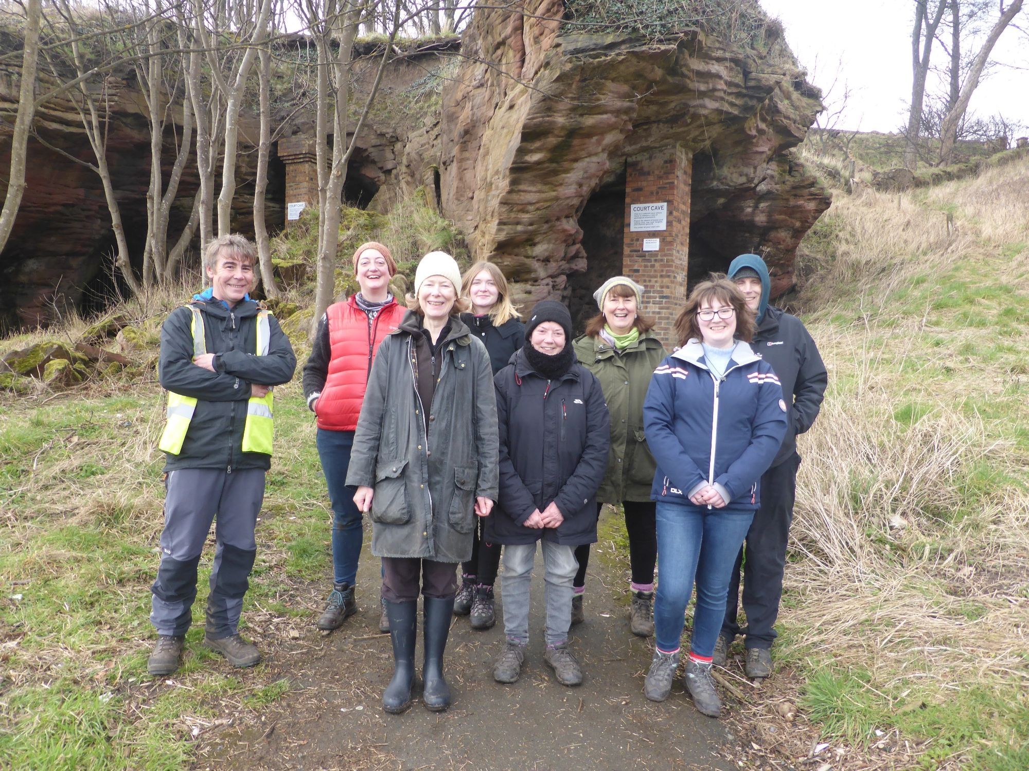 A group of 8 people dressed in outdoor clothing outside a cave.