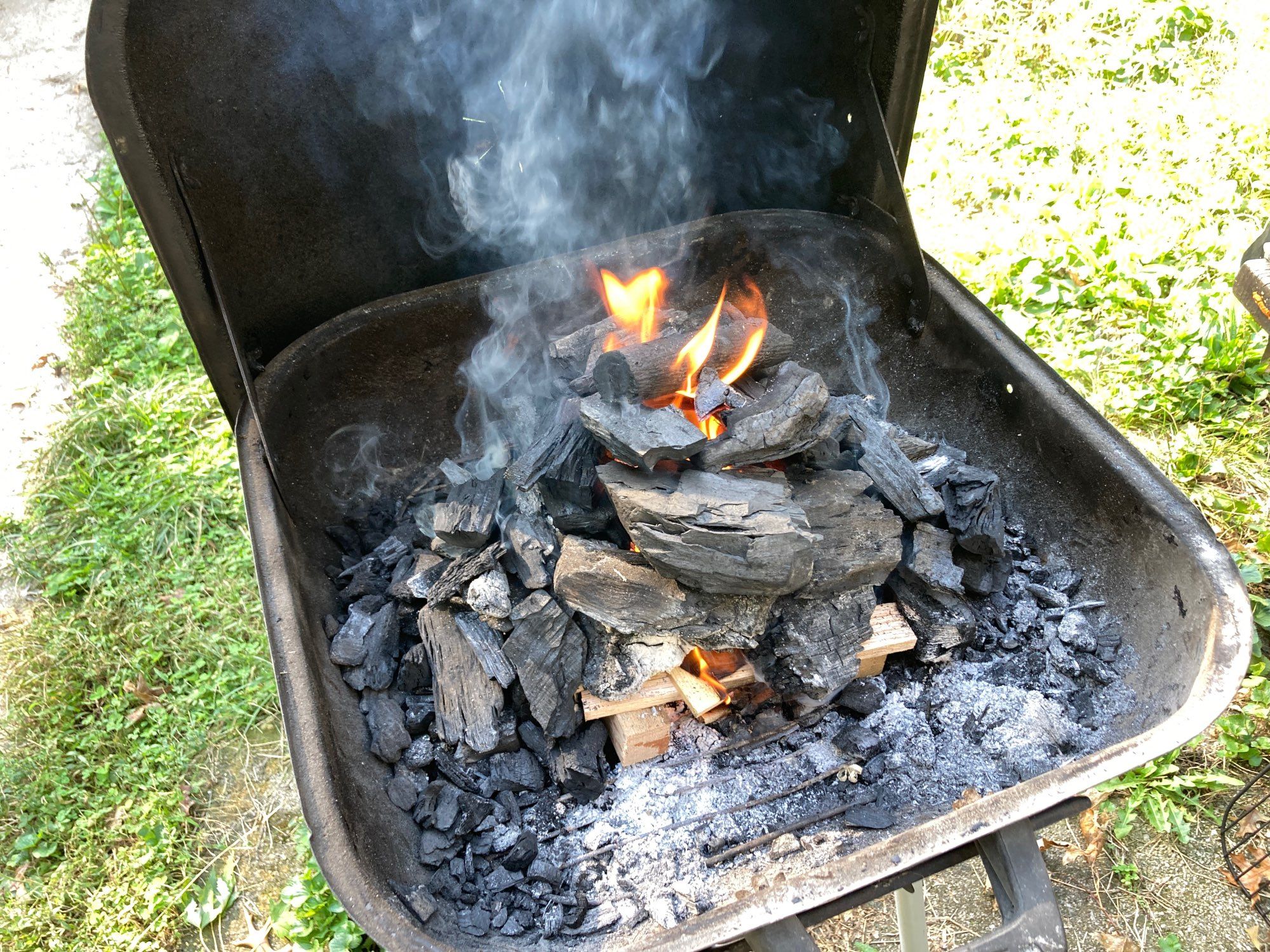 A lump charcoal fire staring in a small, green grill sitting outdoors on green grass on a sunny day. The charcoal is piled in a rough chimney atop a crisscross of hardwood kindling that had been stuff with pine shavings. Flames are spouting out the top.