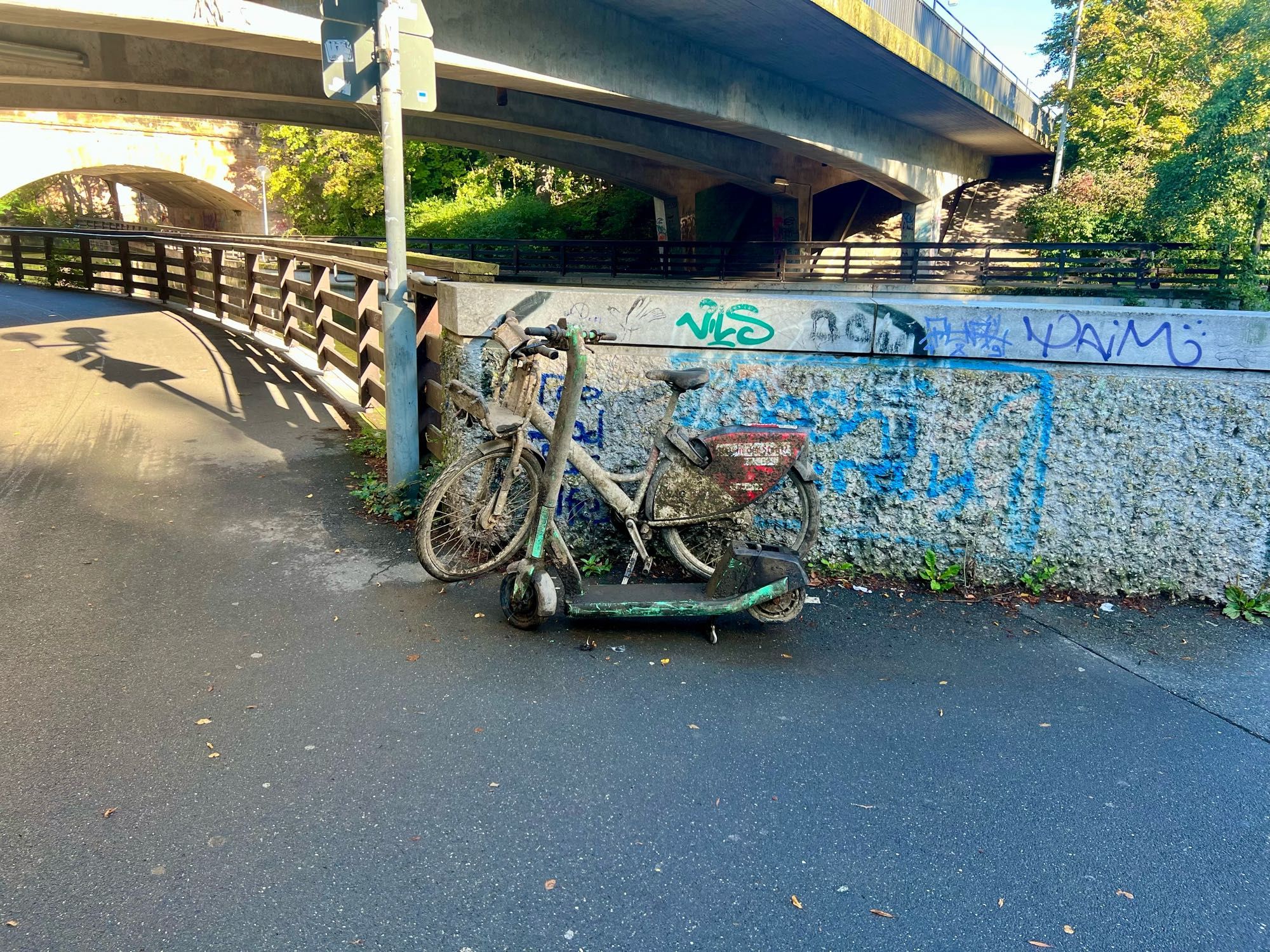 Ein Fahrrad und ein eScooter voller Schlamm stehen neben einer Brüstung am Fluss.