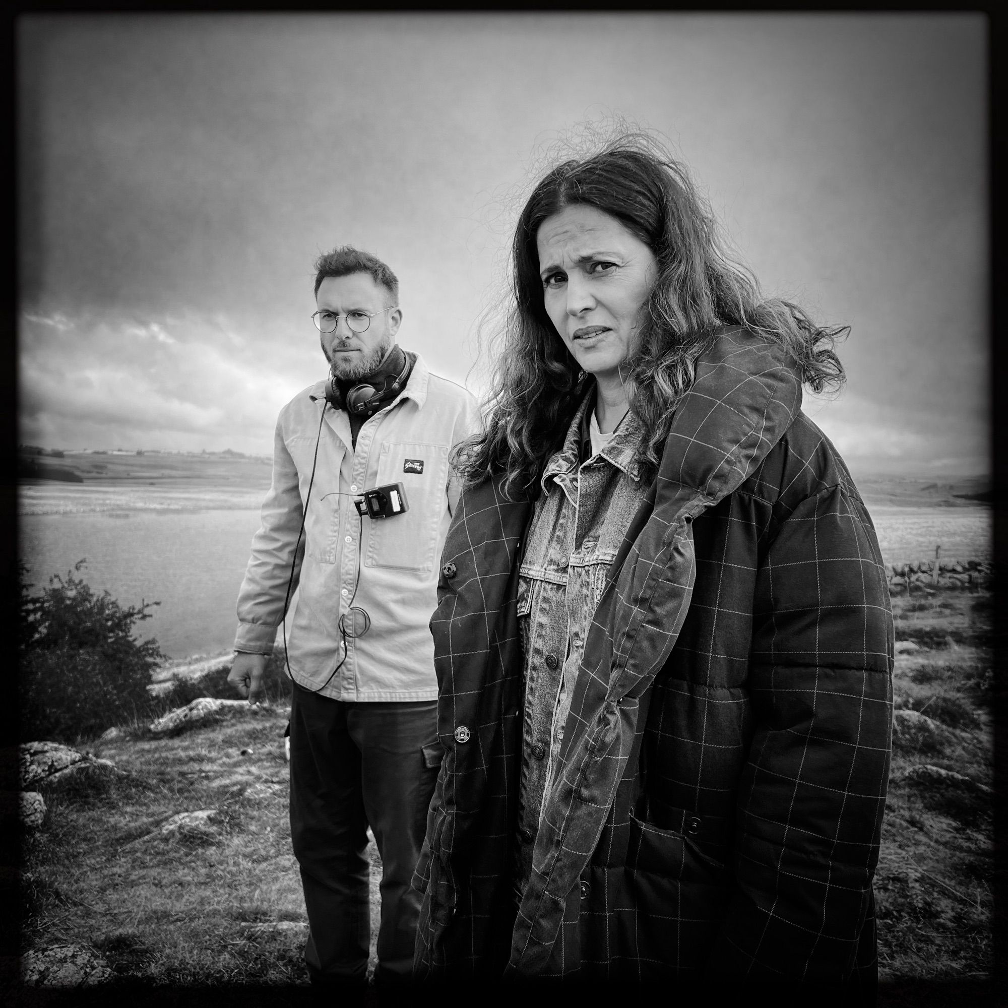 Photo Noir et blanc sur fond d’Aveyron, Franck Brett, réalisateur, l’air concentré, et au premier plan, Anne Charrier, qui nous lance un regard puissant et énigmatique.