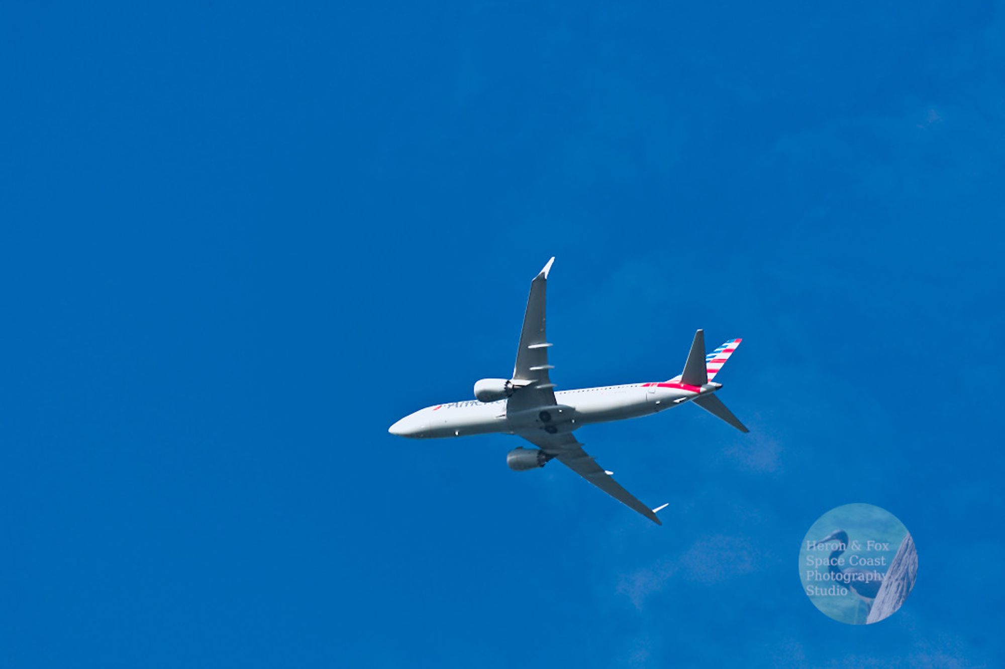 A daylight photograph looking up at an airplane in the sky.
The plane is painted white with the beginning of the word “American” written near the nose and the tail painted with blue and red strips.
The plane has two engines, one of each side. Two landing gear wheels can are nested under the belly of the plane where the wings join.