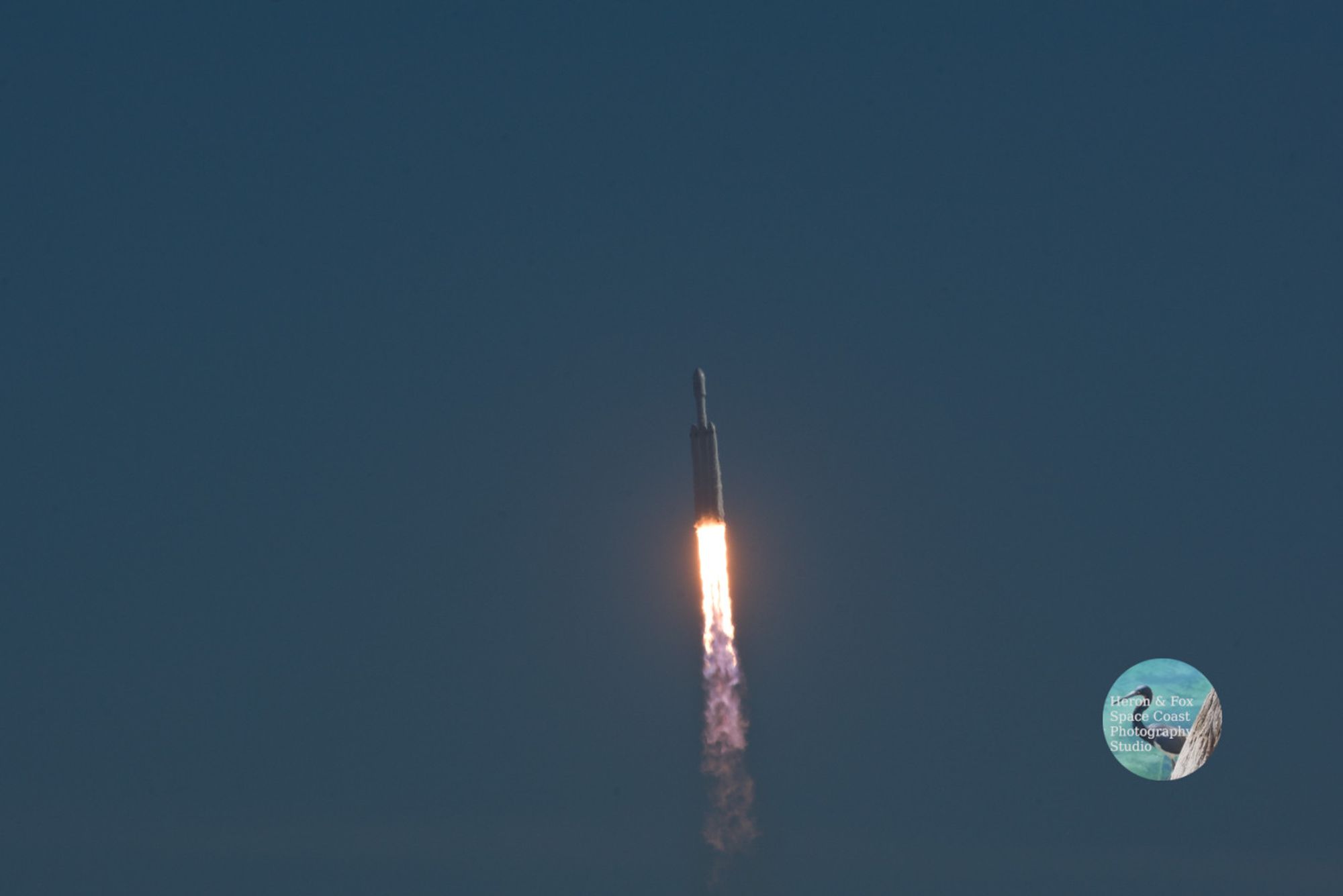 An outdoor, daylight photograph looking at a white rocket against the blue sky.
The rocket is white and has three cylinders on its lower stage with orange flames emerging from the bottom. The center cylinder carries the upper stage and payload fairing.