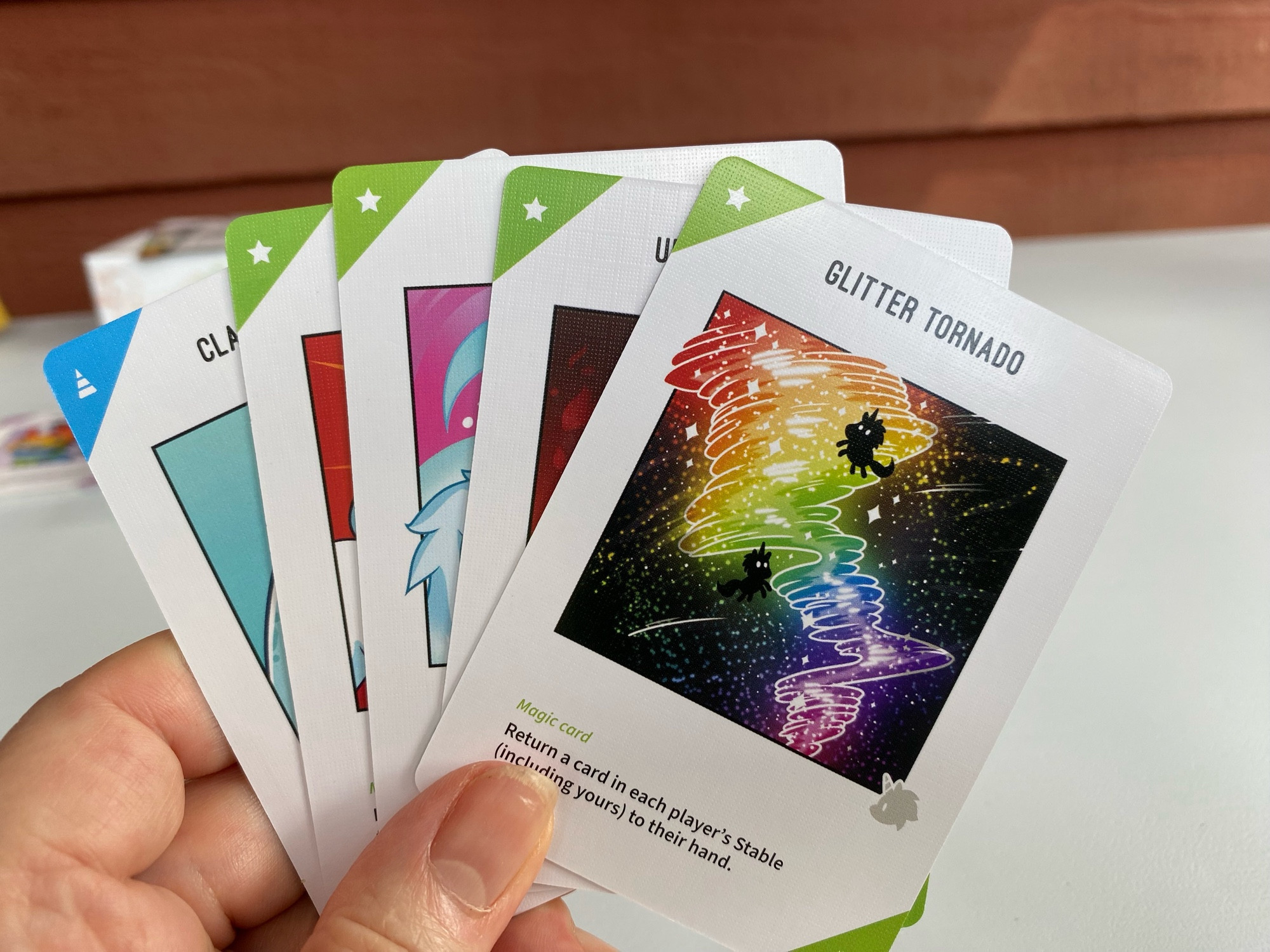 A hand holding five playing cards. The top card is called Glitter Tornado and has a drawing of unicorns floating in a multicolored tornado.