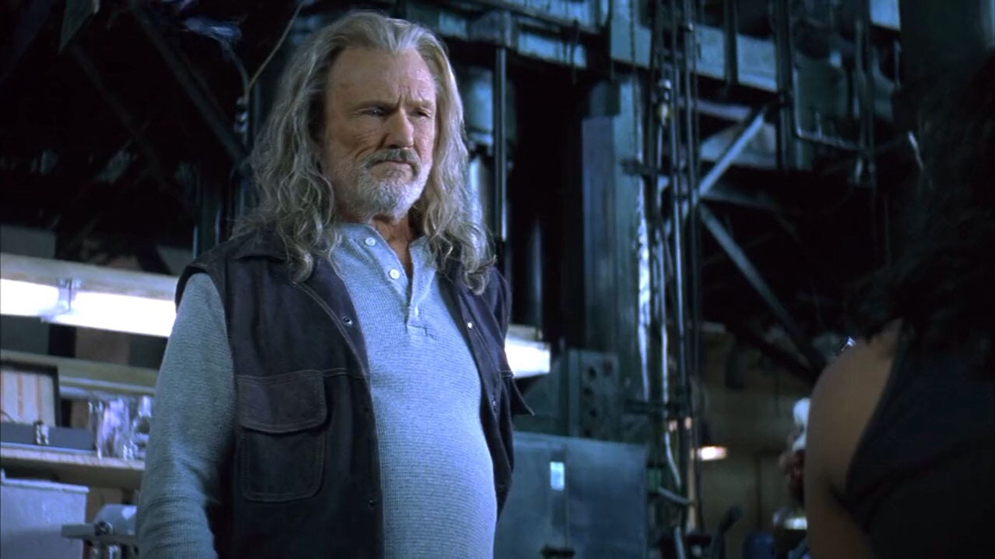 Kris Kristofferson in Blade. He's inside a dark warehouse, wearing functional, workmanlike clothing: a long sleeve shirt and either demin or leather vest. He has long grey hair and a scruffy beard.