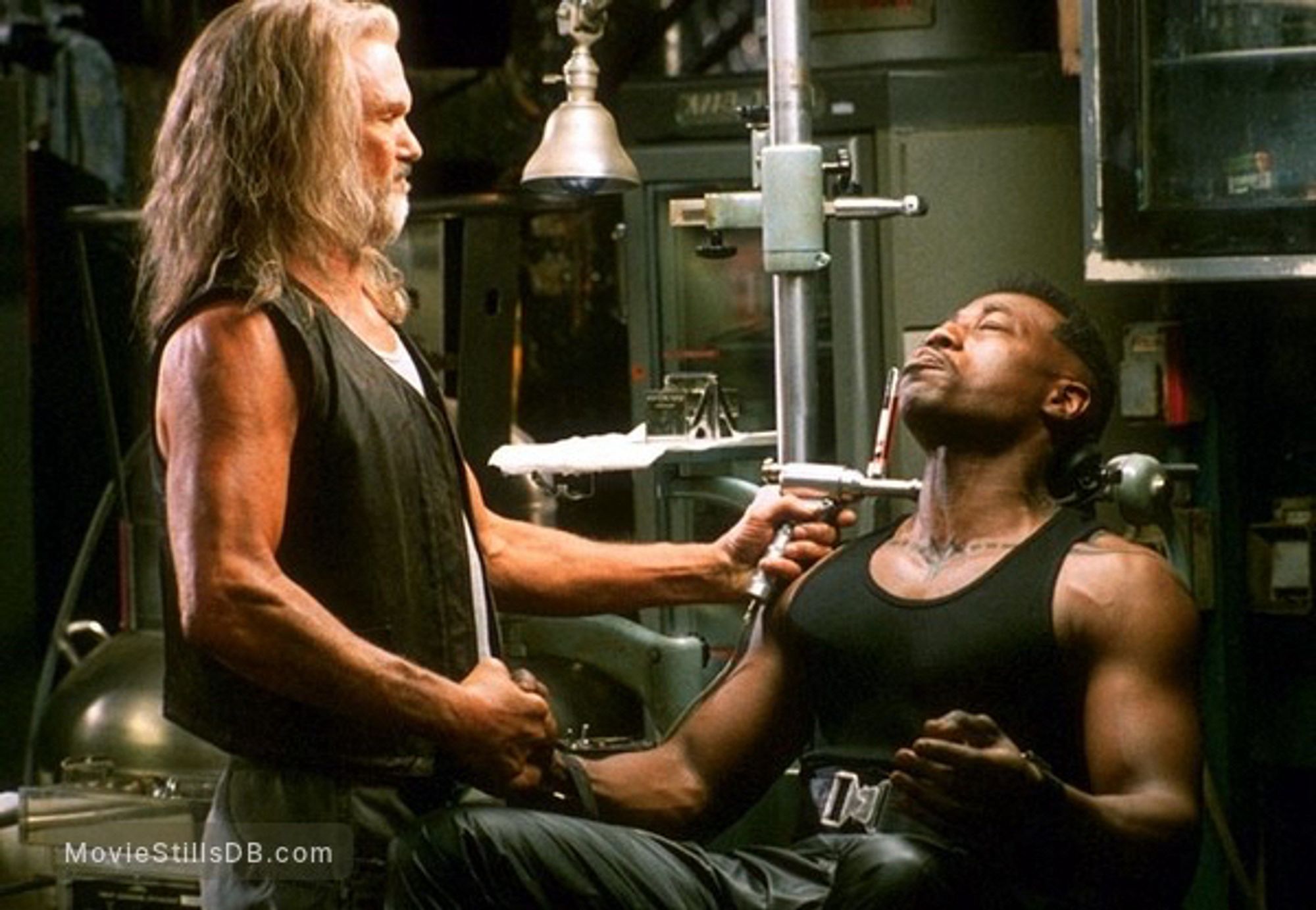 Kris Kristofferson and Wesley Snipes in Blade. They are inside darkened laboratory. Both are wearing black tank tops. Snipes is seated and Kristofferson is injecting him in the neck. They are holding right hands.