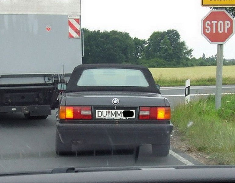 Ein altes BMW Cabrio von hinten an einem Stop-Schild. Sein Kennzeichen DU MM, was sich also dumm liest. 