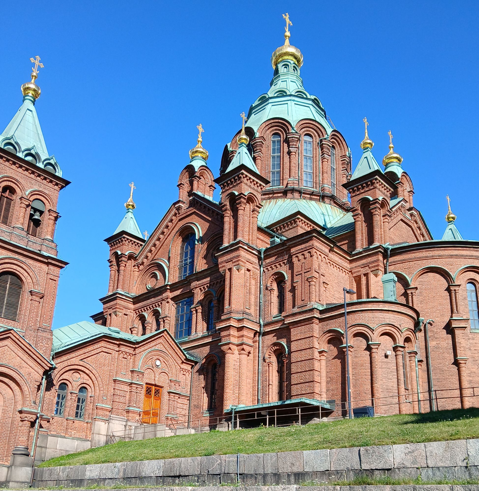 prächtige russisch-orthodoxe Kirche in historisierend-byzantinischem Stil mit hohen Rundbögen, zahlreichen Türmchen und goldfarbenen Dachverzierungen - ansonsten Dächer "kupfergrün", Mauerwerk blassrot vor blauem Sommerhimmel (Die Kirche steht auf einer Anhöhe.)