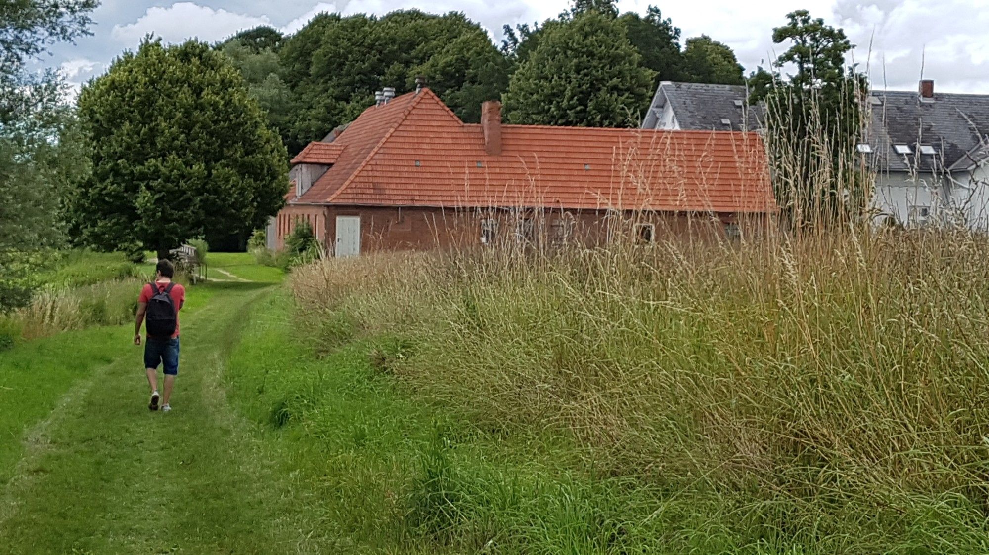 Junger Mann mit rotem Shirt, Jeans-Shorts und schwarzem Rucksack geht auf einem grasbewachsenen Feldweg auf einen Bauernhof zu, der aus roten Ziegeln mit ebenfalls rotem Ziegeldach errichtet ist.