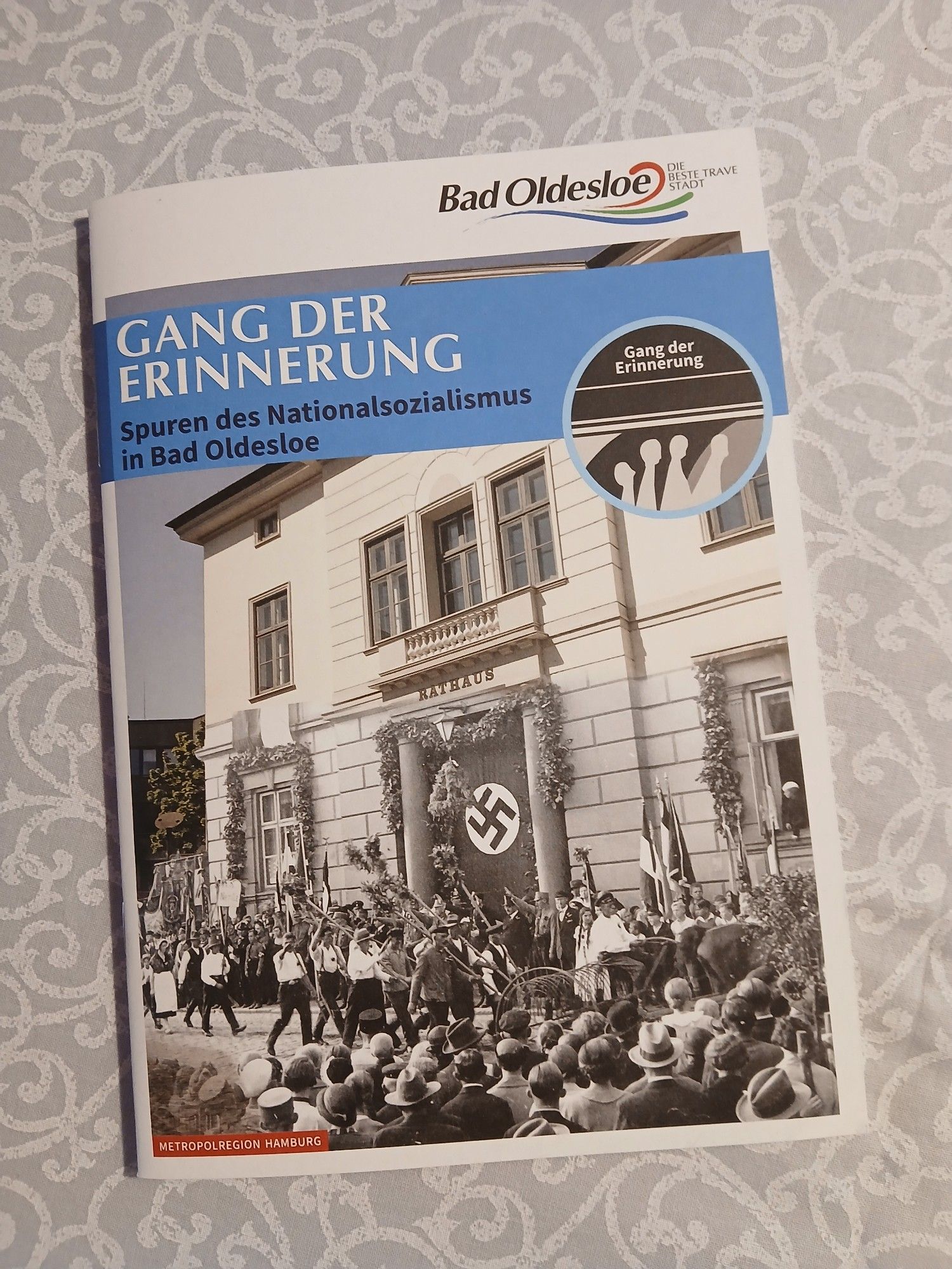 Broschüre "Gang der Erinnerung - Spuren des Nationalsozialismus in Bad Oldesloe" mit dem offiziellen Logo der Stadt, einen runden Erkennungsmotiv "Todesmarsch" und einen ganzseitigen Umschlagfoto, das aus zwei Ansichten des alten Rathauses zusammengesetzt ist: der obere Teil aus heutiger Zeit und in Farbe, der untere mit einer Hakenkreuz-Fahne und einer Ährenkrone in der Tür und einem vorbeiziehenden NS-"Erntedank"-Aufmarsch.