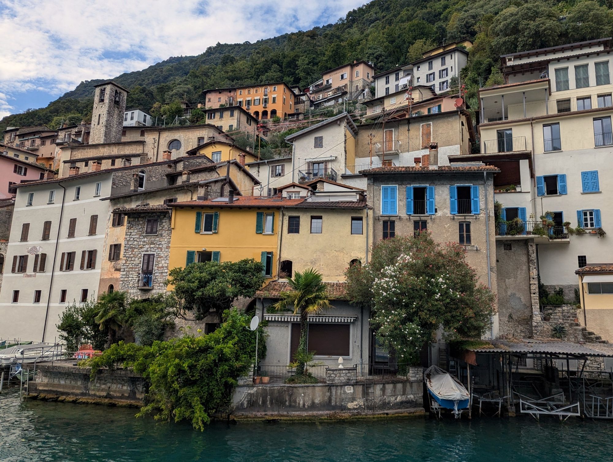 A small terraced village with colorful houses on Lake Lugano, seen from the lake looking up the hill.