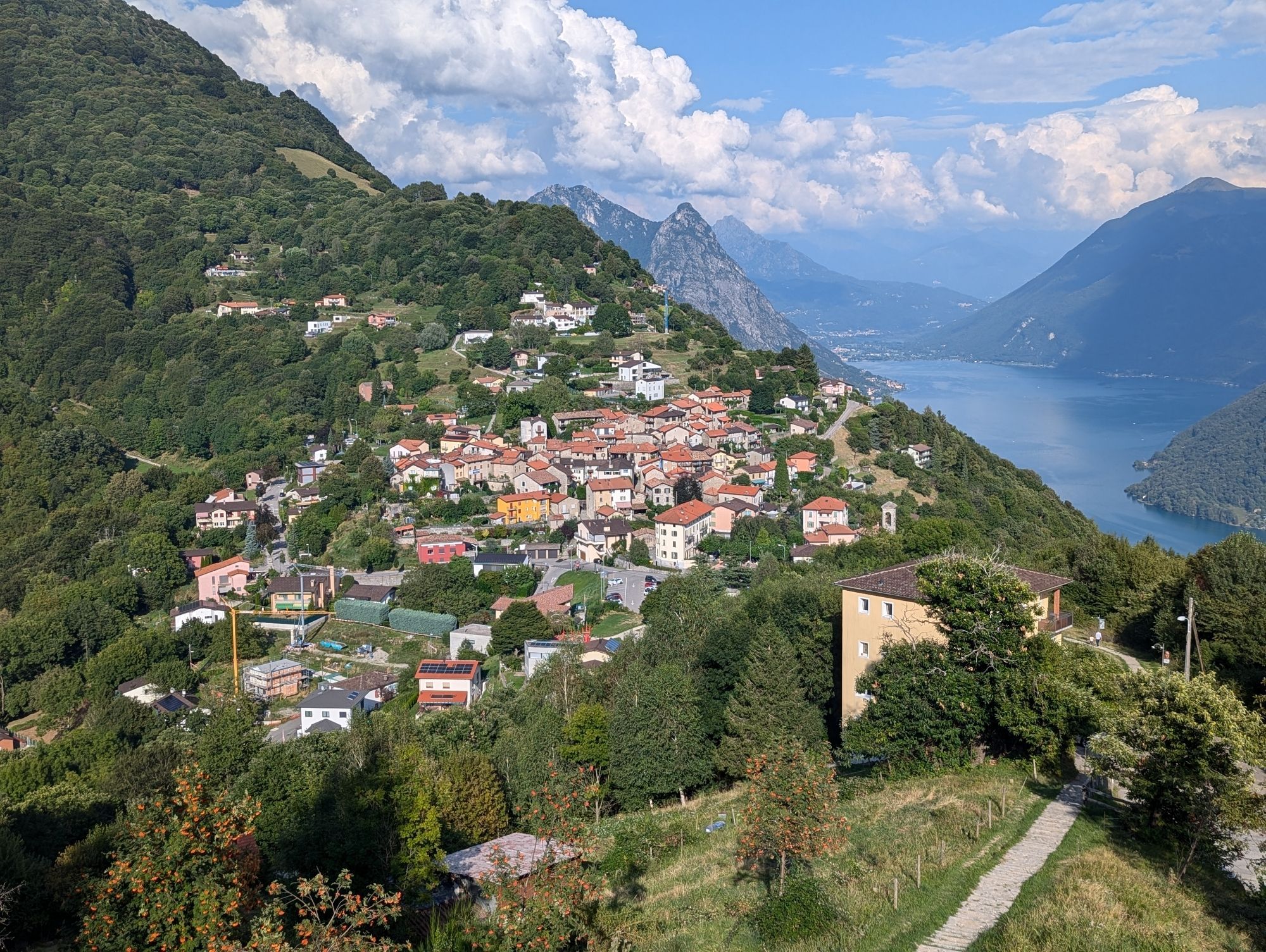 In Alpine surroundings the image is looking over a small village on the side of a hill. It is surrounded by forest. Downhill to the right is a blue lake surrounded by steep hills and mountains.