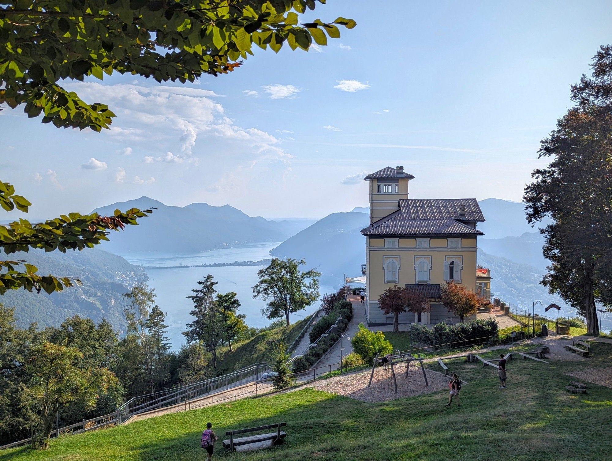 A solitary house stands on a vantage point overlooking alpine mountains surrounding a lake that stretches out into the distance.