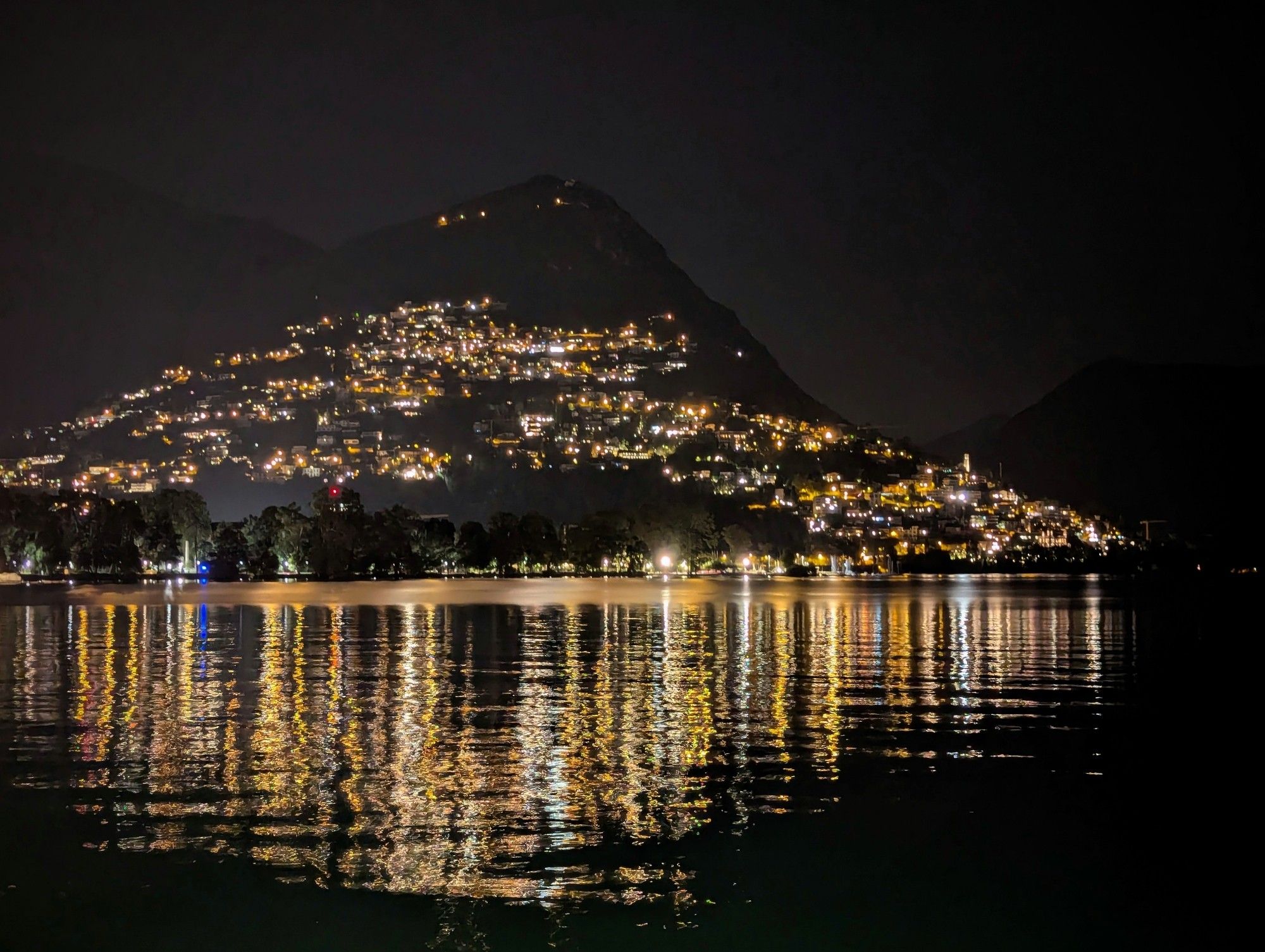 A night time image taken of a village on the side of the hill, in the foreground is a lake and the lights from the village are reflected in the surface of the lake.