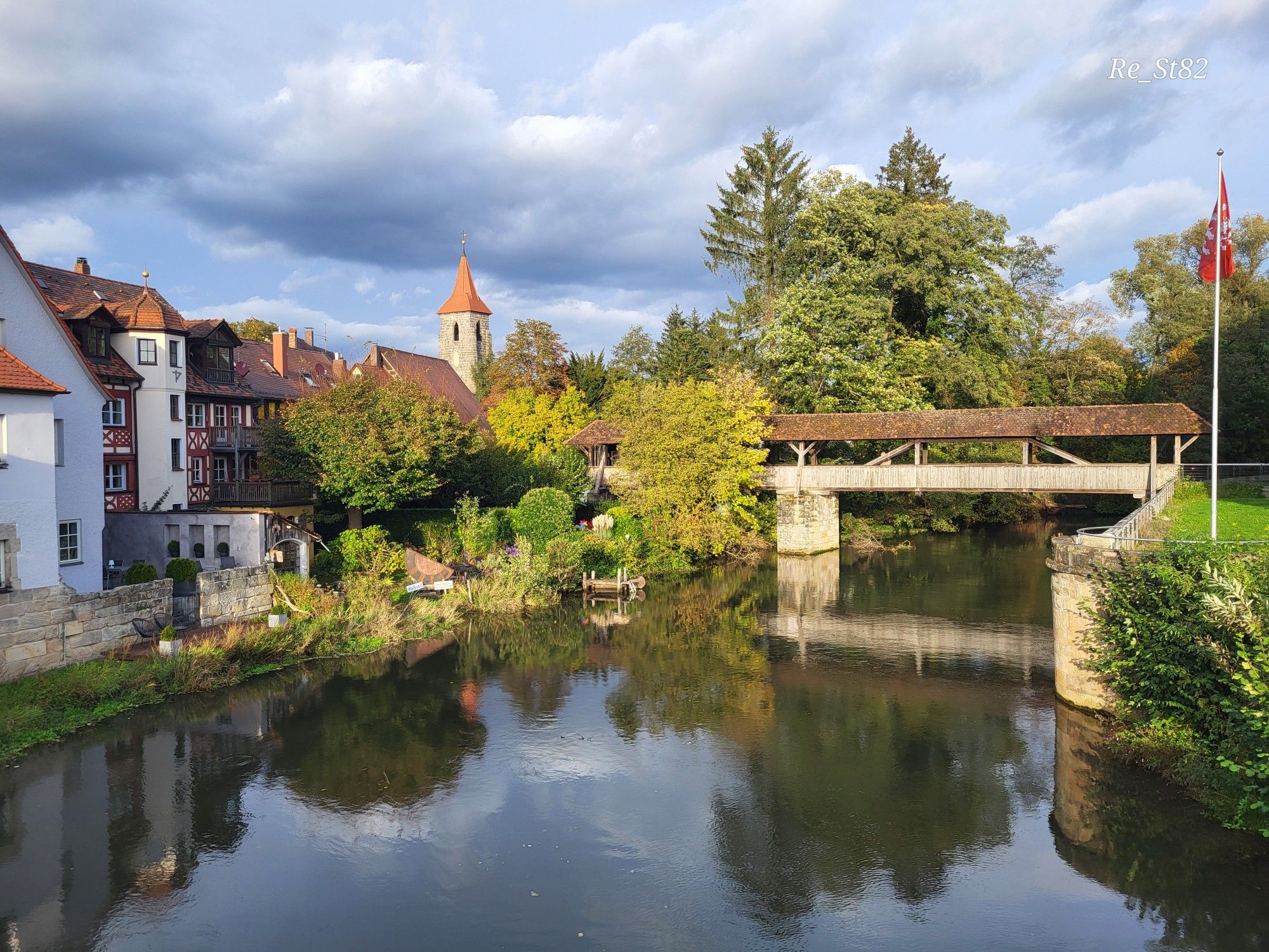 Links niedrige Wohnhäuser, vor diesen Bäume und Wiese an einem Fluss. Über den Fluss führt eine überdachte Fußgängerbrücke die scheinbar an einer Stadtmauer endet. Der Himmel ist bewölkt, aber die Sonne scheint. 