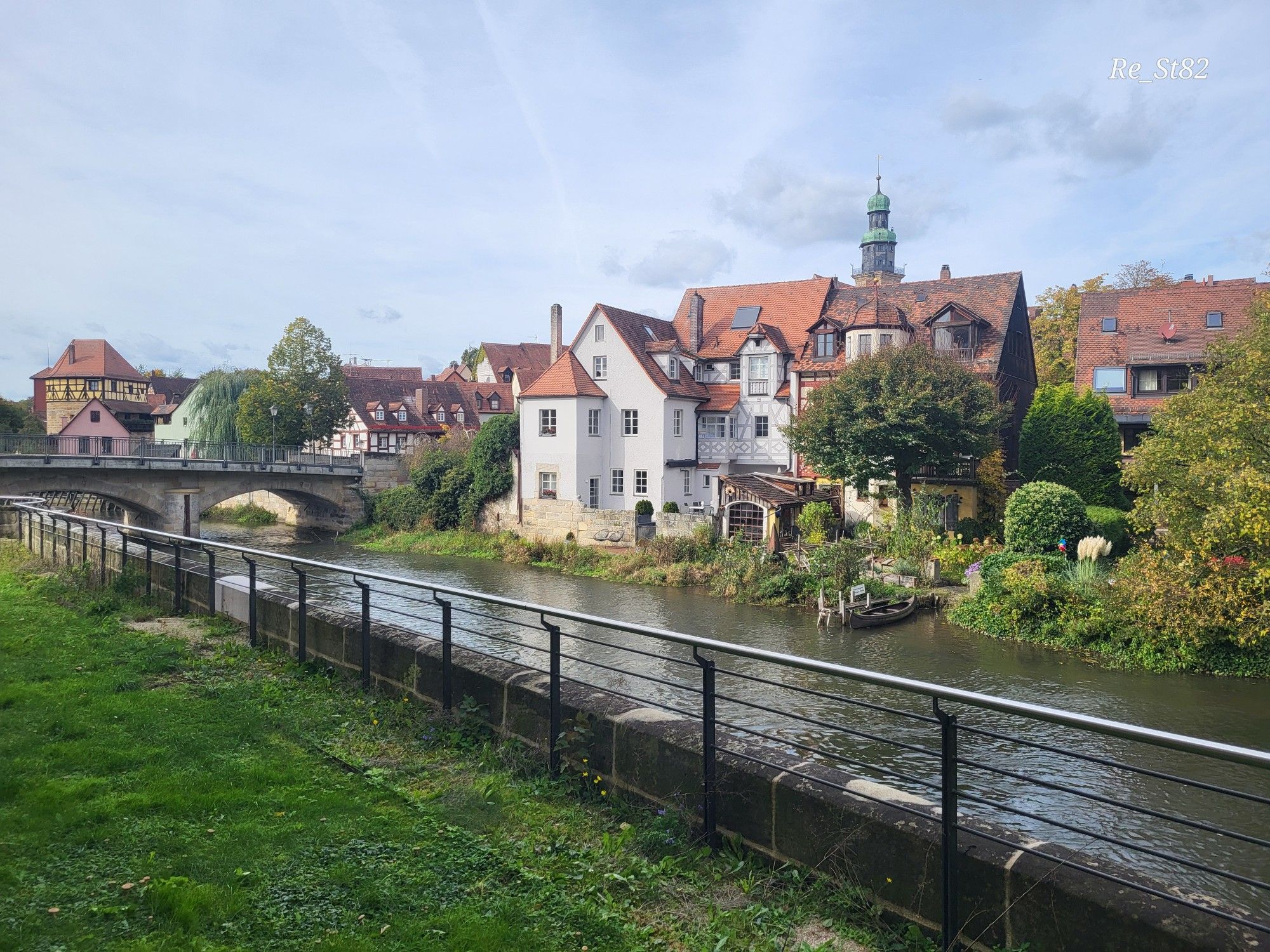 Wiese, links hinten eine Brücke über einen Fluss, auf der anderen Seite des Flusses Wohnhäuser, im Hintergrund ein Kirchturm, das Flussufer ist grün, an einem Holzsteg liegt ein Ruderboot. 