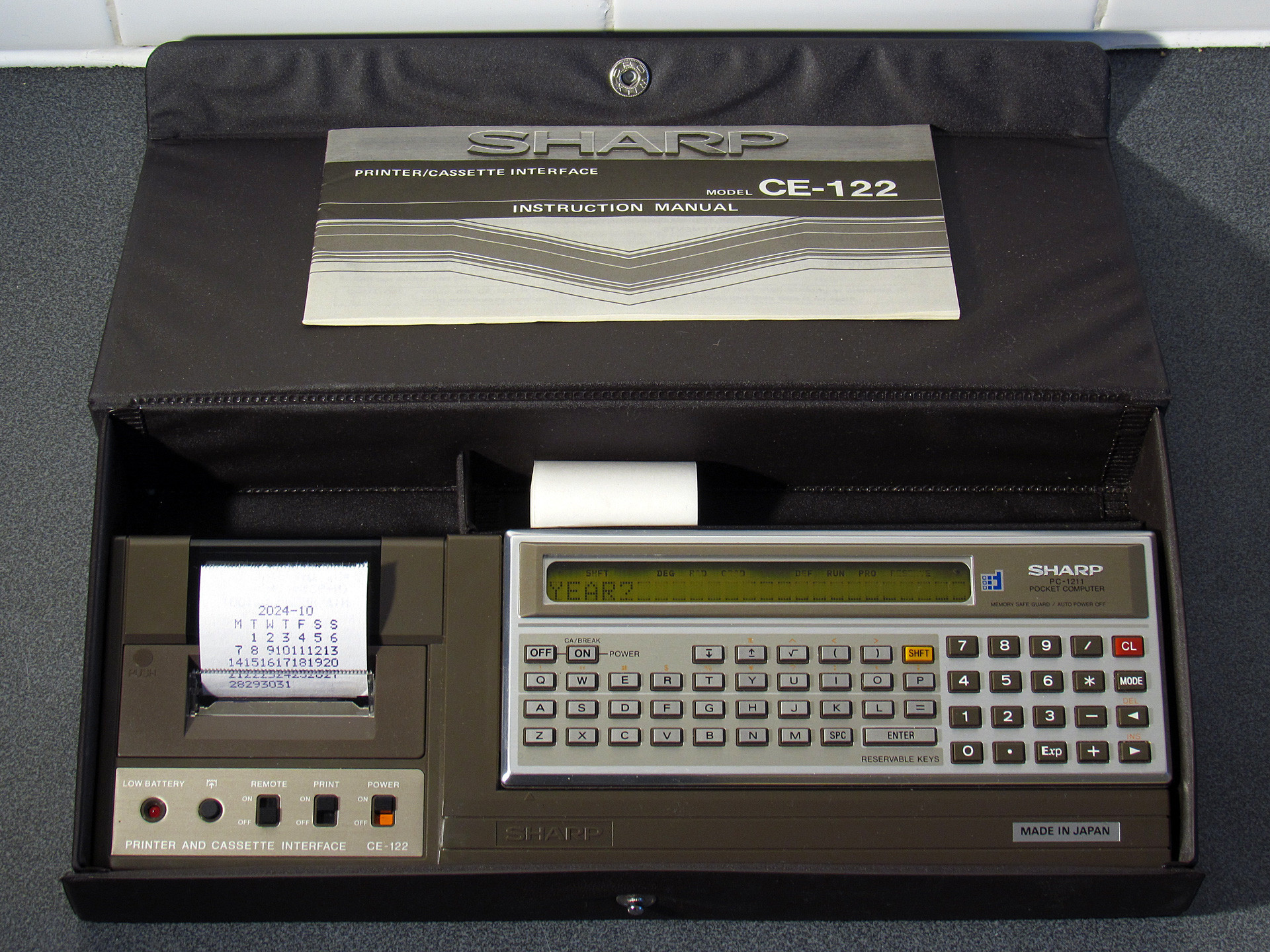 Photo of Sharp PC-1211 connected to the CE-122 printer and cassette interface, showing a monthly calendar printed by the computer.