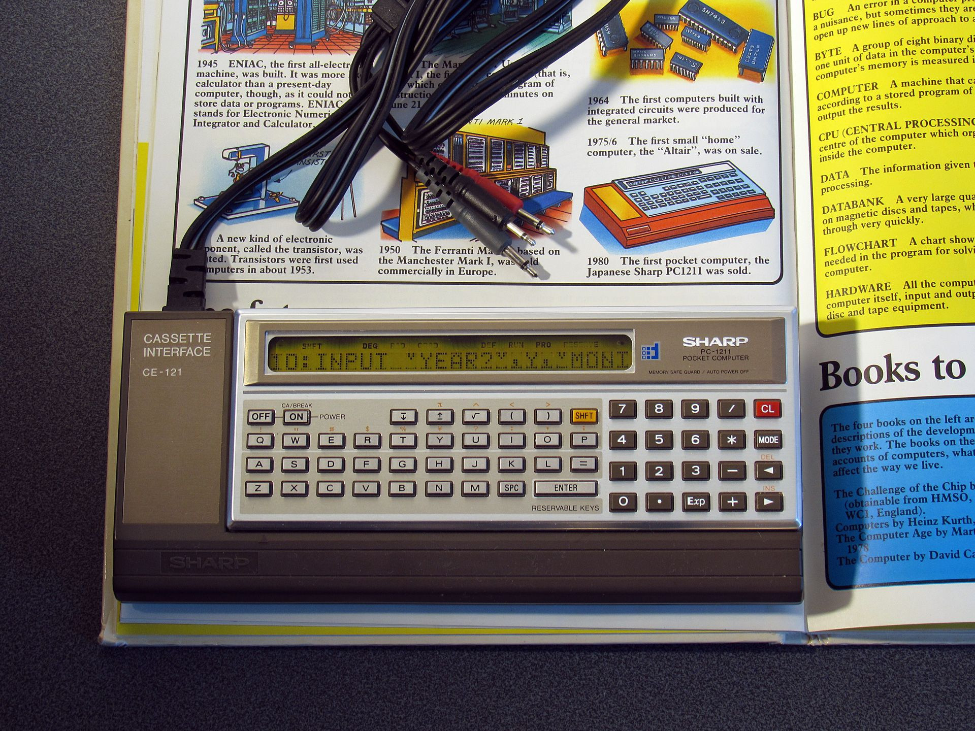 Photo of a page describing the Sharp PC-1211 as "the first pocket computer" with the PC-1211 inside the CE-121 cassette interface on top.