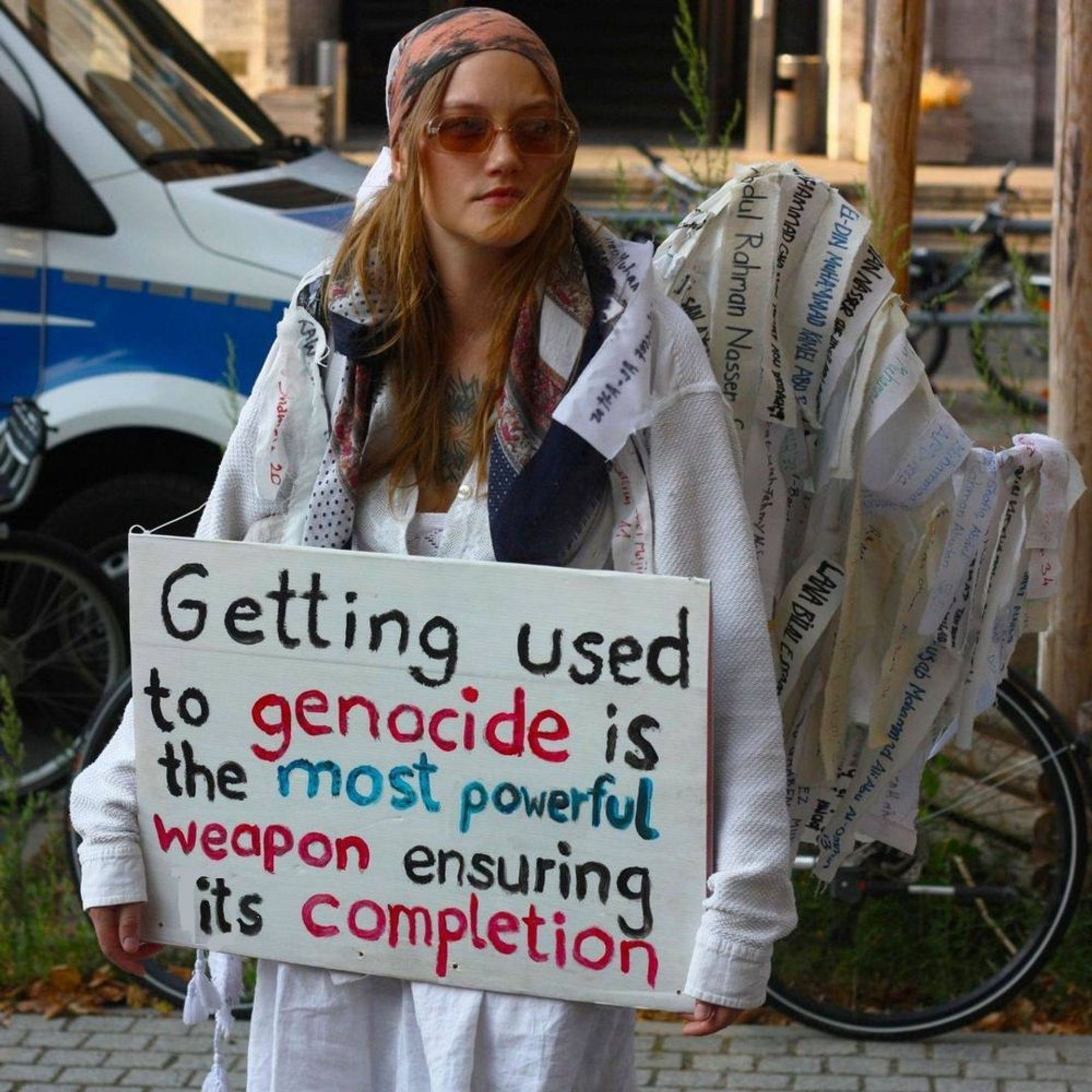 A Berlín demonstator holds a sign which reads "Getting used to genocide is the most powerful weapon ensuring its completion".

Behind her, a bicycle is covered in strips, each with the name of a murdered Palestinian. The woman is wearing one such strip around her shoulders.