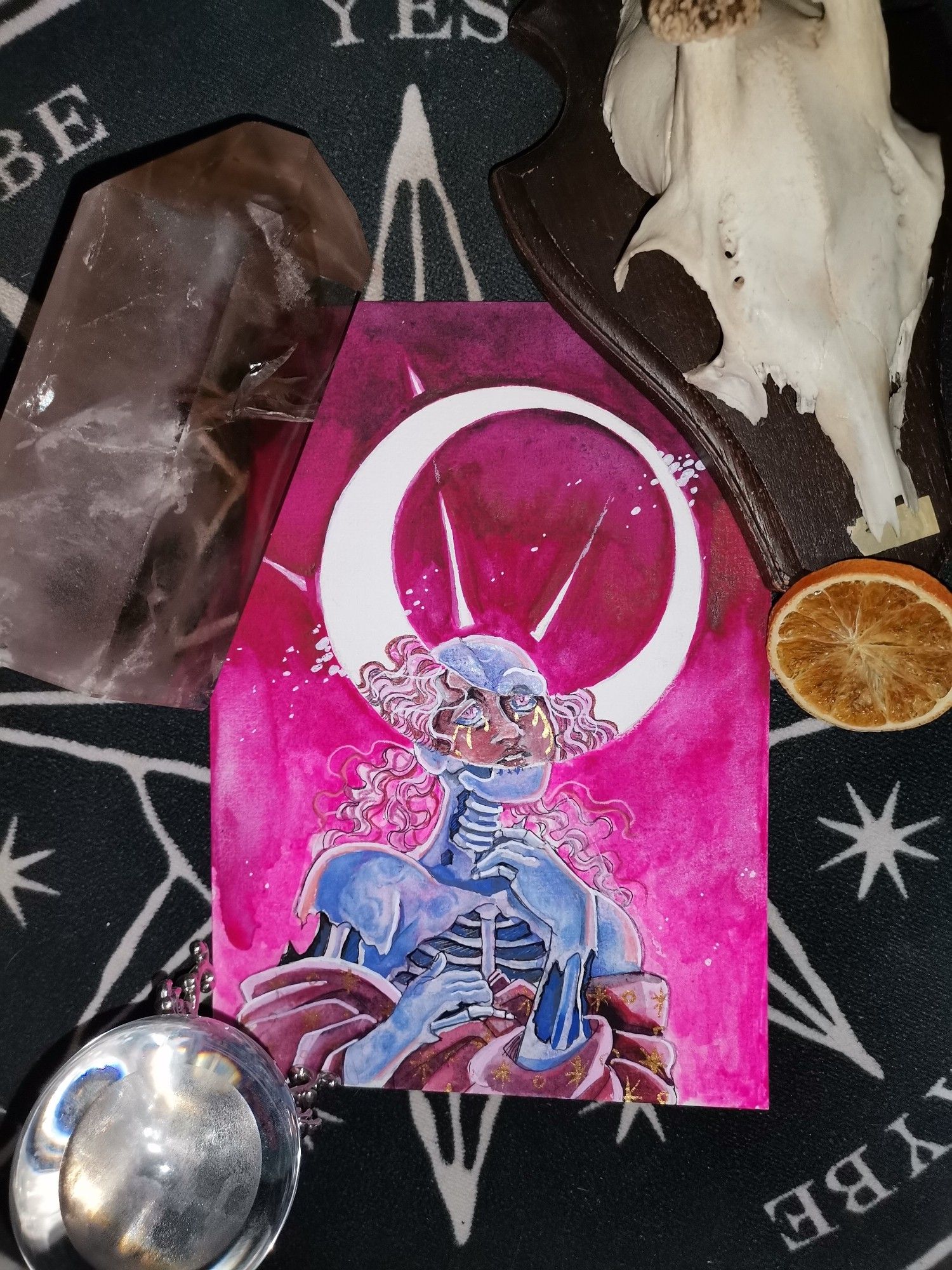 Image of a person who seems to be a decaying blueish corpse. Part of their face is covered by a crescent moon, where they seem alive and well, but crying and looking up. The image is surrounded by a smokey quartz, a chrystal ball with the moon in it, the skull of a deer and a piece of dried orange.