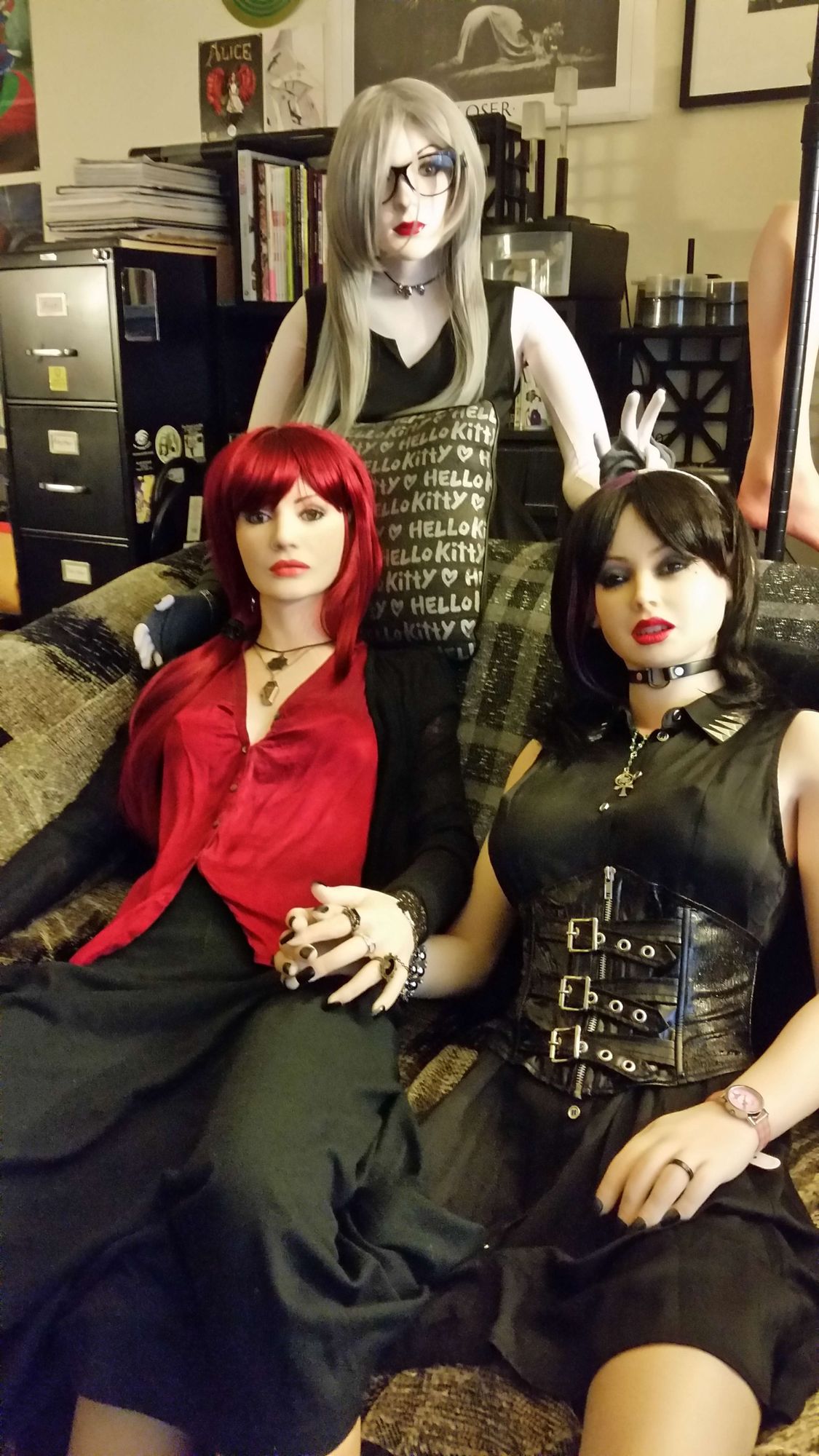 From left to right: Elena Vostrikova, a companion Doll made by Anatomical Doll, Muriel Noonan, a companion Doll made by Textile Doll, and Sidore Kuroneko, a RealDoll by Abyss creations. Elena and Sidore are seated on a brownish-grey loveseat and holding hands, while Muriel is standing behind them, making bunny ears with her right hand behind Sidore's head. All three are stylish Synthetik Goth women, and are making direct eye contact with the viewer.