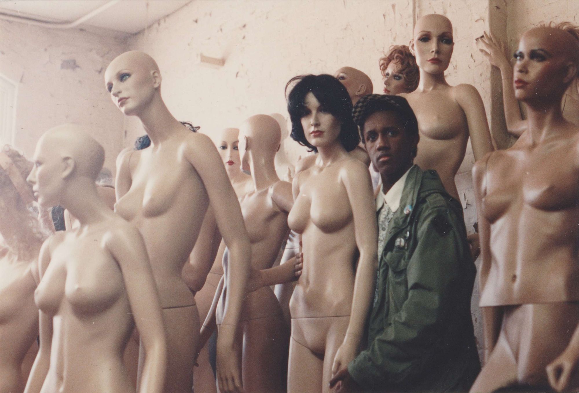 Davecat is wearing a black baseball cap with a black-and-white chequered interior, a green US army parka, a white dress shirt with a black pattern, and is making direct eye contact with the viewer as he stands with his arms around the waist of a naked mannequin woman with medium-length black hair. They are surrounded by a crowd of naked mannequin women of various skintones