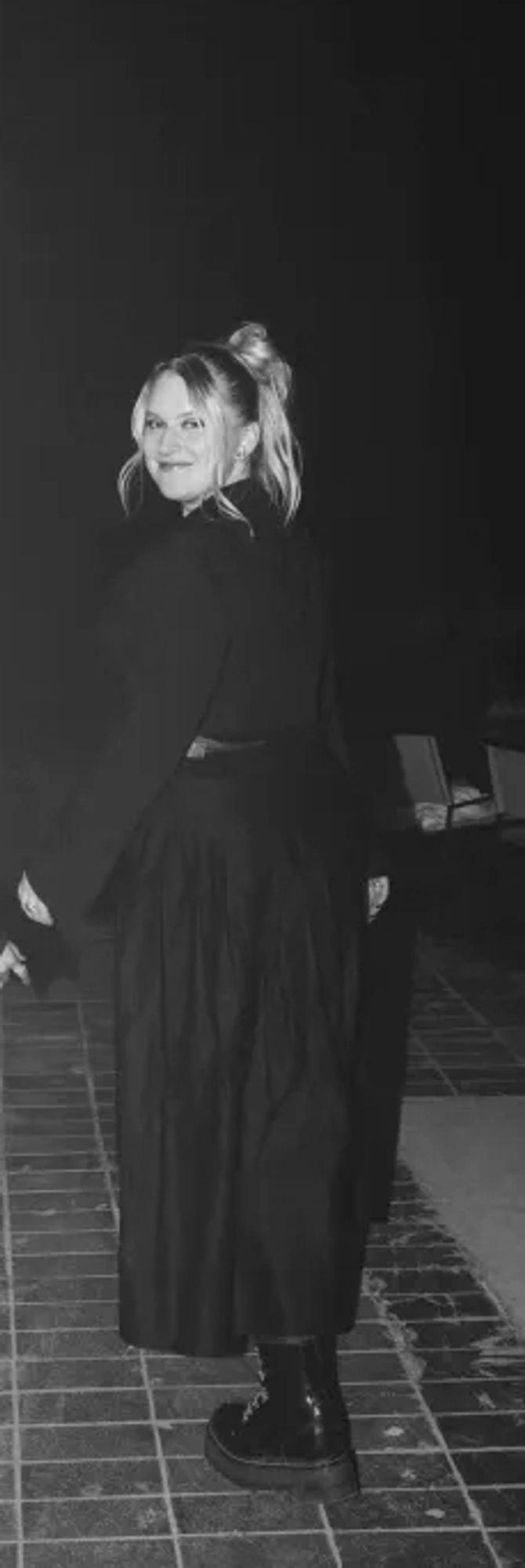 Matilda Belin Larsson in a blonde person in a black and white photo. She's walking down a brick-paved street at night wearing black lace-up boots, a calf-length black skirt, and a black jacket.