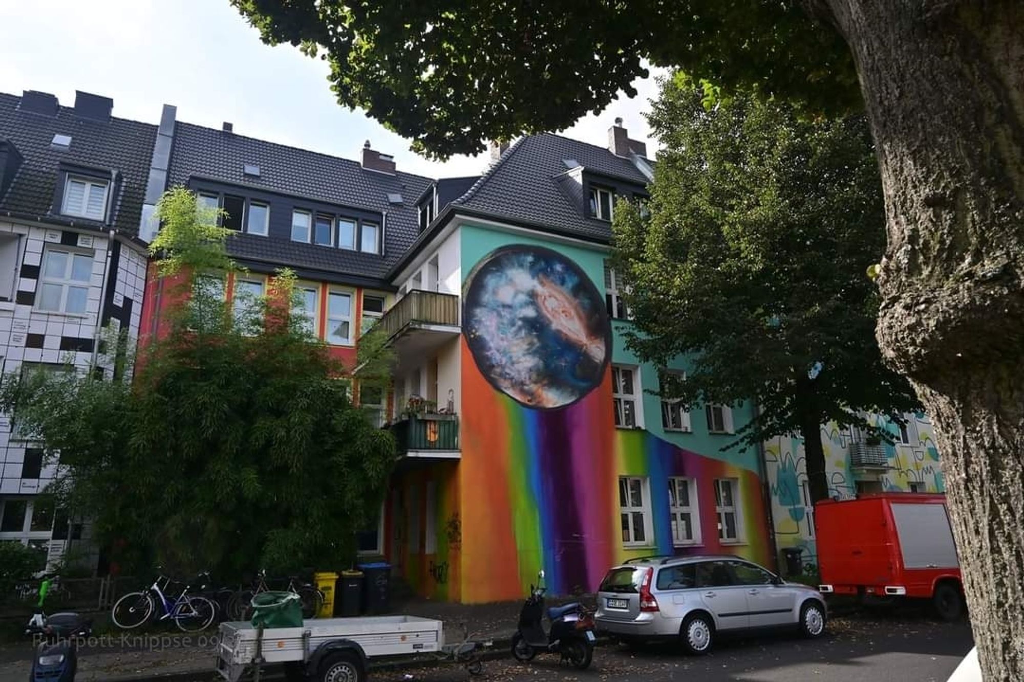 Eine in Regenbogenfarben bemalte Fasseäade eines Wohnhauses, im oberen Teil schwebt eine Weltkugel, die eine Galaxie in sich zeigt.