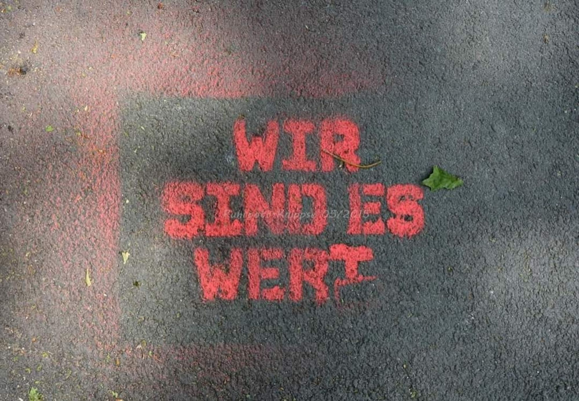 Ein auf Betonboden in roter Farbe aufgesprühter Schriftzug: 
" WIR
SIND ES
WERT"