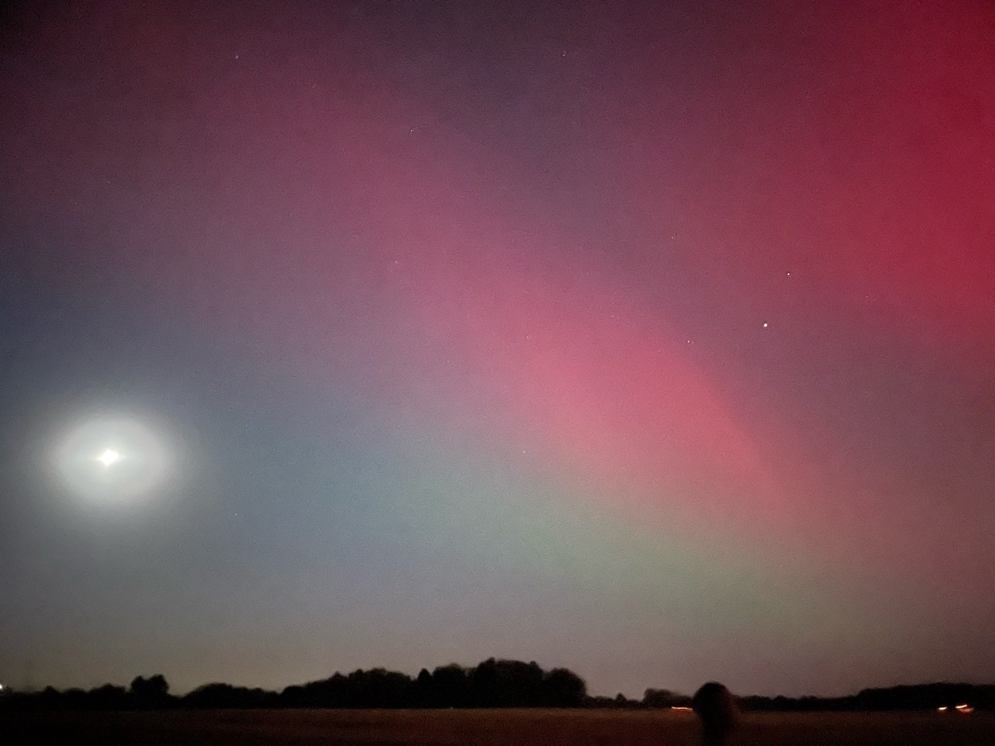 Green and red aurora borealis streaks the night sky with the hazy moon off to one side