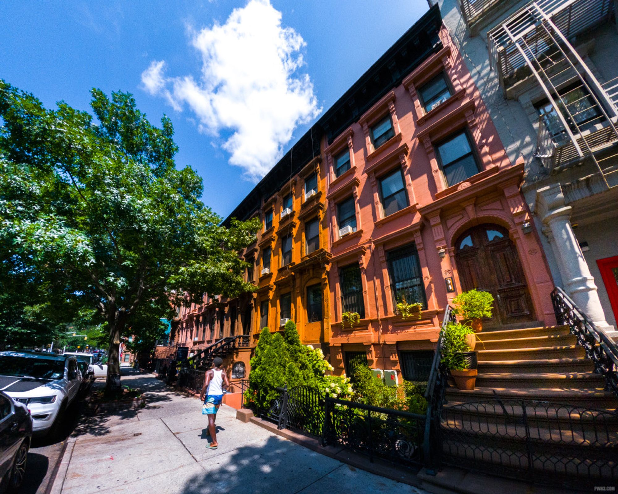 We are on 124th street in Harlem, facing west. A young African-American boy wearing blue swimming shorts and a white tank top walks along the street. He is walking on the sidewalk in front of a row of colorful brownstone homes. The street is lined with trees and a few parked cars. The sky is blue with a cloud in the center.