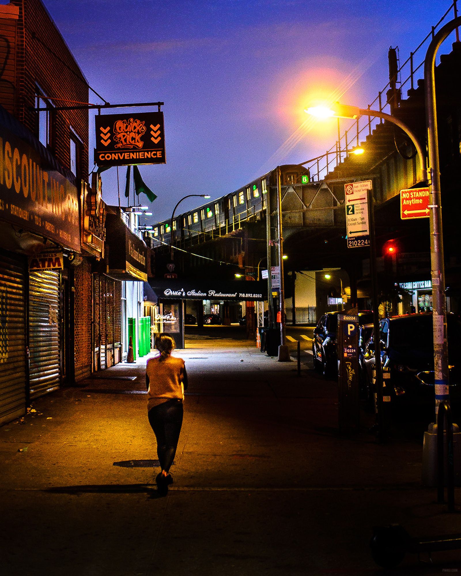 A photograph of a woman walking along a sidewalk with a subway train in the distance. We are on a sidewalk in The Bronx, New York City. It's early morning and still dark but the sky is starting to get lighter. As we look down the sidewalk we see a lone woman in a light-colored sweater and black pants walking away from us. On the left side of the sidewalk there are many closed stores, including one with a sign that says "Quick Pick Convenience". On the right side of the sidewalk we see the elevated train tracks of the Number 6 subway train. In the distance a 6 Train moves along the elevated tracks, starting to curve around us.