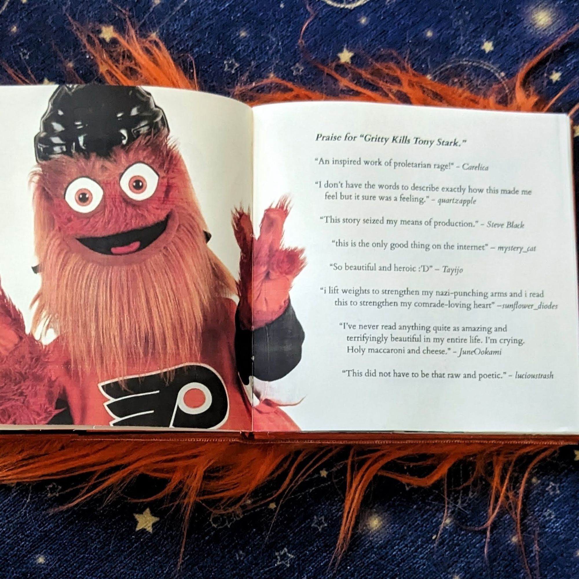 A spread from the fuzzy orange book. At left, Gritty makes a peace sign. At right are a series of quotes (from commenters on the AO3 fic) under the heading “Praise for Gritty Kills Tony Stark.” Quotes include “An inspired work of proletarian rage” and “this story seized my means of production”