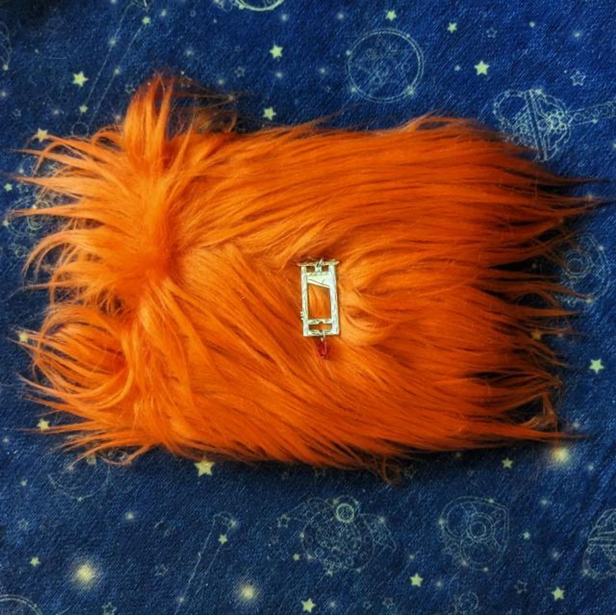 A book bound in ORANGE FUR with a gold guillotine pin. It looks like a malevolent orange tribble, but you want to touch it. It is sitting on a piece of blue fabric with a subtle print of stars.