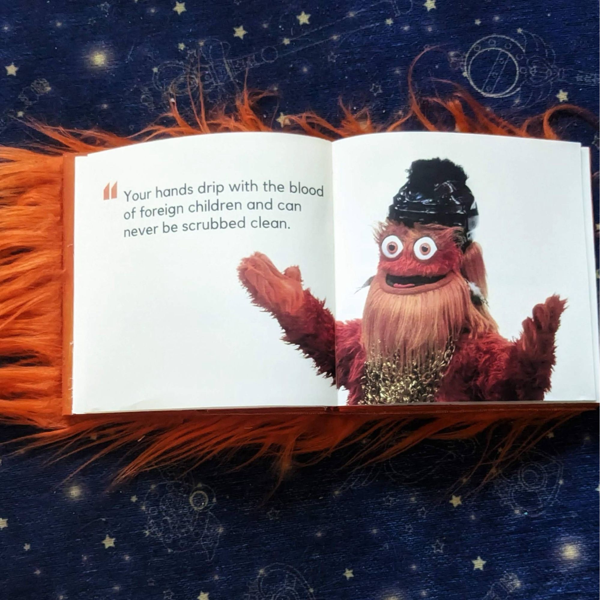 Another spread from the fuzzy orange book, Gritty Kills Tony Stark: a picture of Gritty (orange fuzzy hockey mascot) spreading his arms. Text next to him says “Your hands drip with the blood of foreign children and can never be scrubbed clean.”