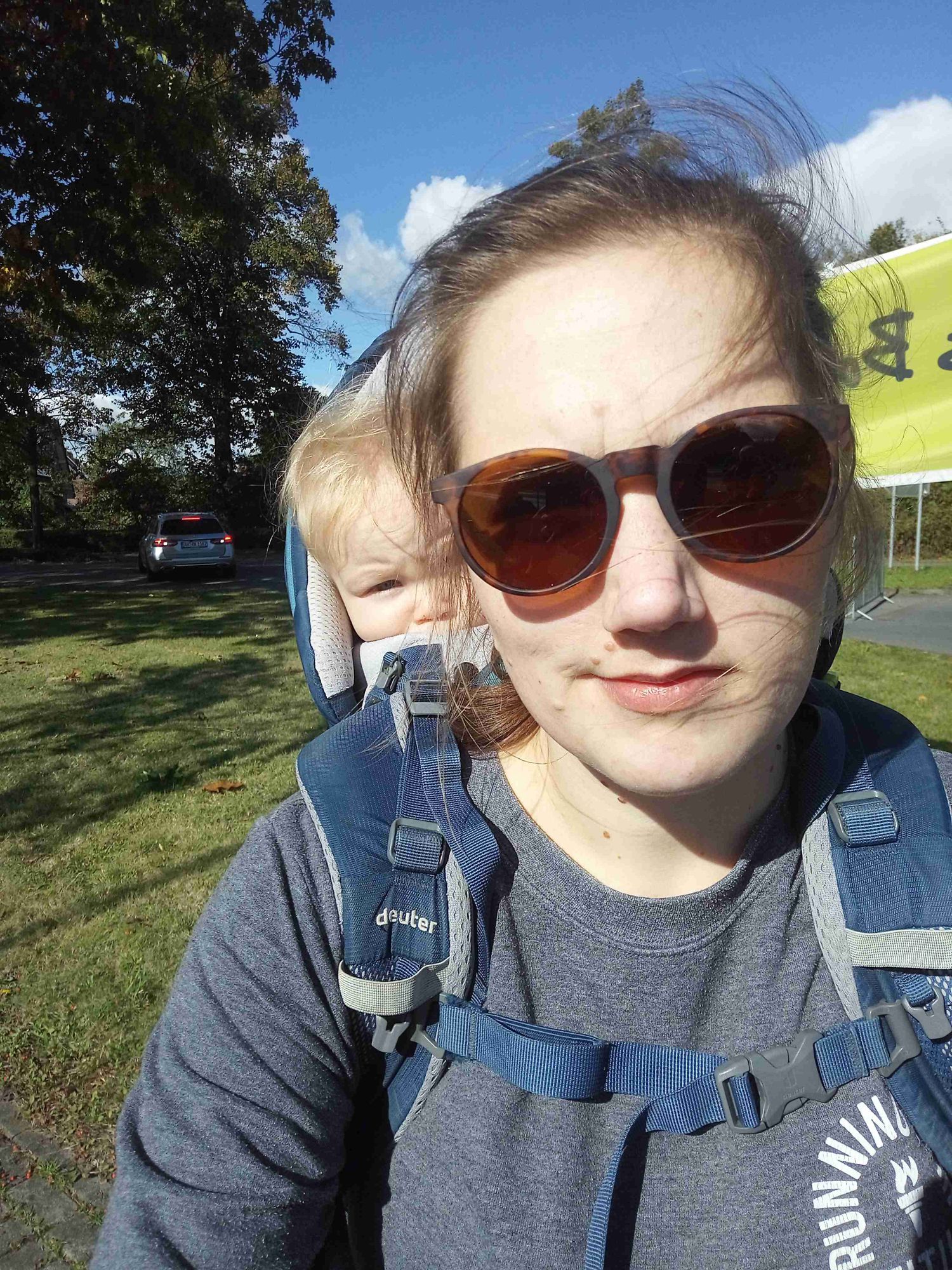 Selfie, Frau mit Sonnenbrille und Kleinkind in einer blauen Kraxe auf dem Rücken