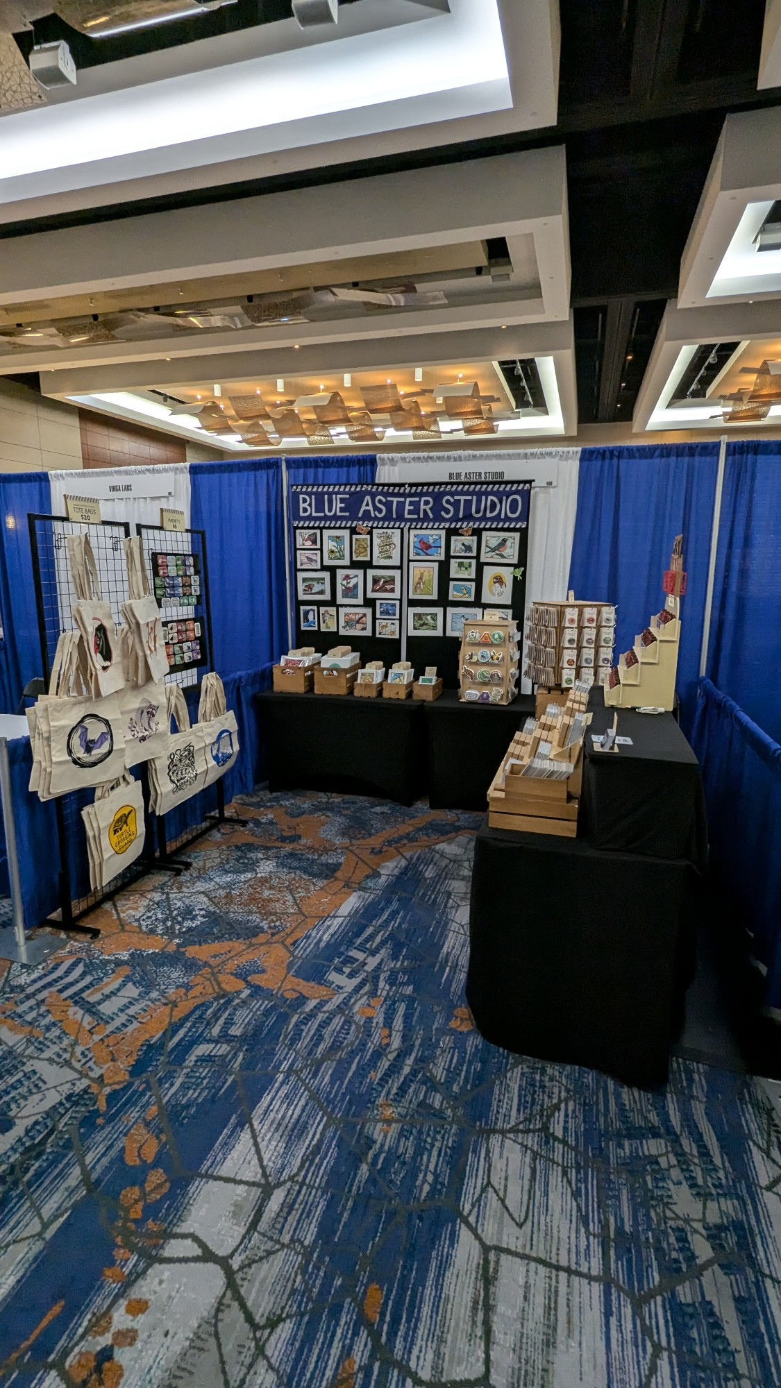 The blue Aster Studio booth all set up with wildlife inspired art, tote bags, magnets, stickers, ornaments, stationery and buttons