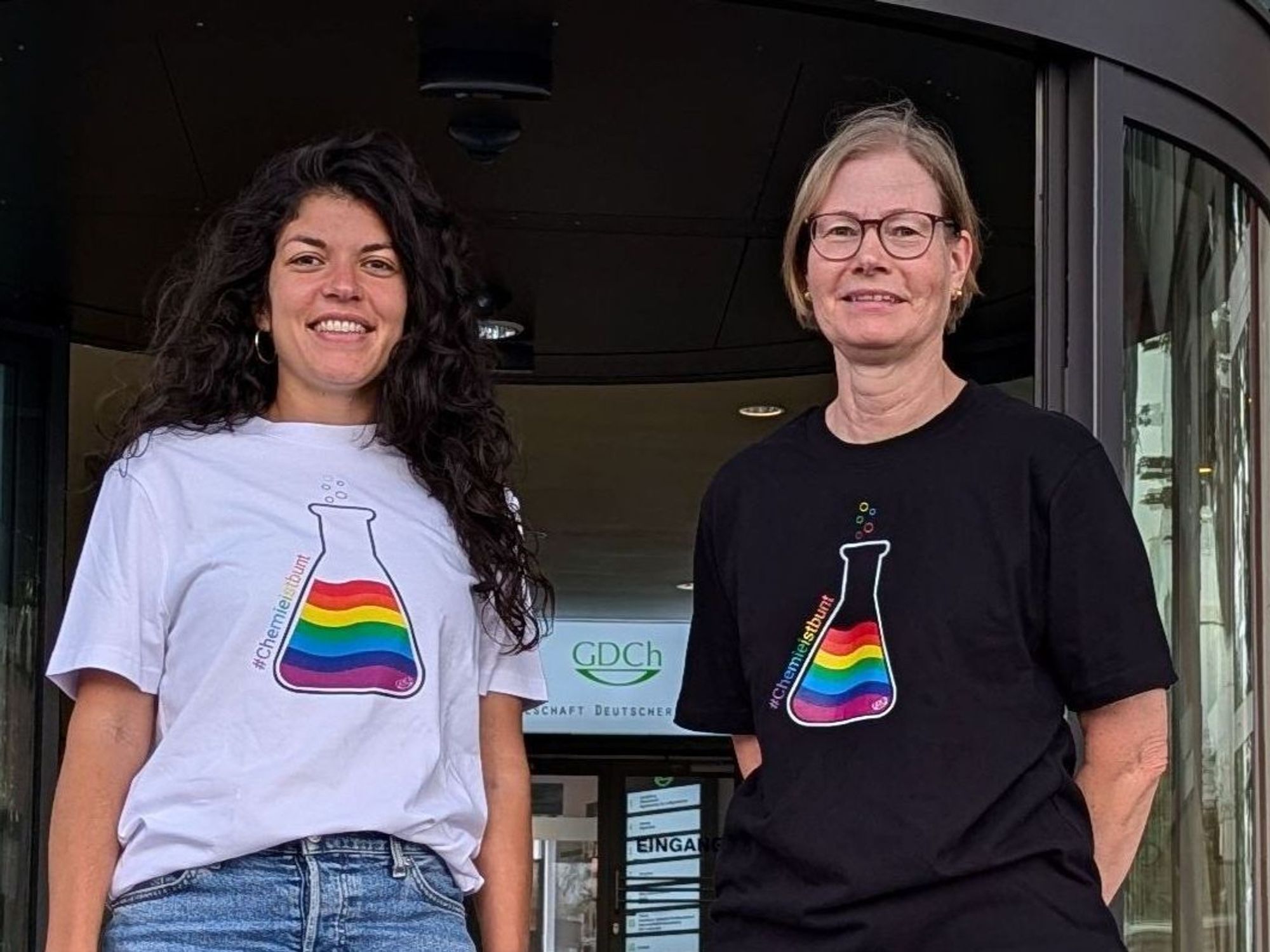 Zwei Frauen tragen je ein T-Shirt mit einem Erlenmeyer-Kolben, in dem sich eine Flüssigkeit in den sieben Regenbogenfarben befindet.