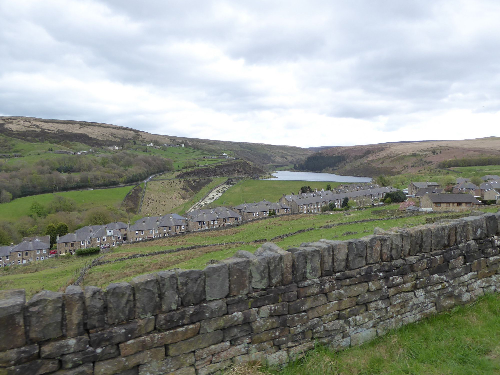 Butterley Reservoir nestled in the Wessenden valley, brown moorland above, and lush green grass on the dam embankment. Dark stone houses and dry stone walls line the roads.