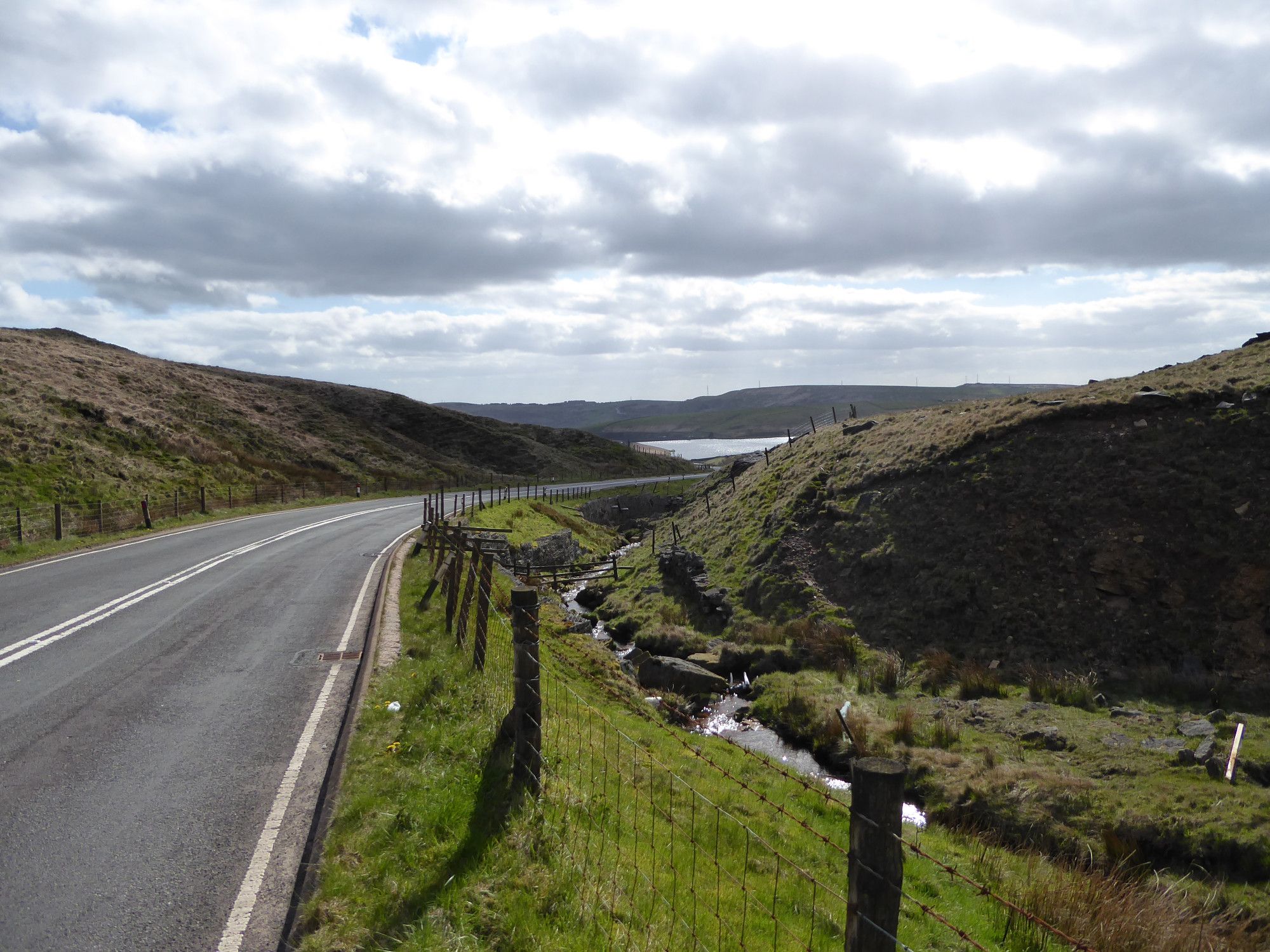 Looking back towards Dowry Reservoir, the sunlight bright in the clouds and sparkling on the road and water surfaces. The road descends toward a corner and disappears towards the reservoir.