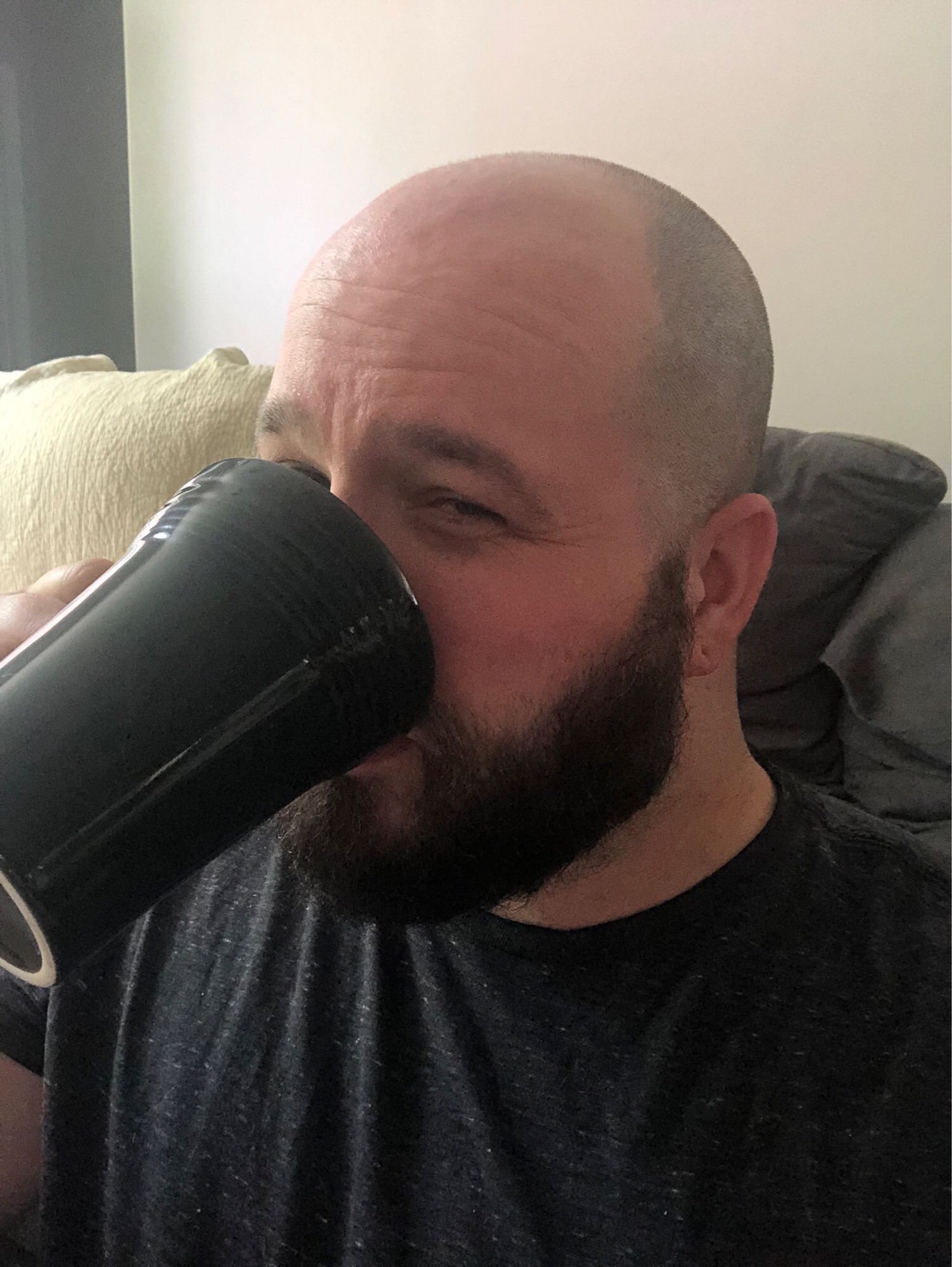 Brettybooop sipping on coffee out of a large black ceramic mug. He is bald. He is still wearing the textured black shirt. His beard is still freshly trimmed. Outside of the boundaries of this image resides the chaos of a child’s toys strewn across the landscape. Interior shot. iPhone 10 quality image.