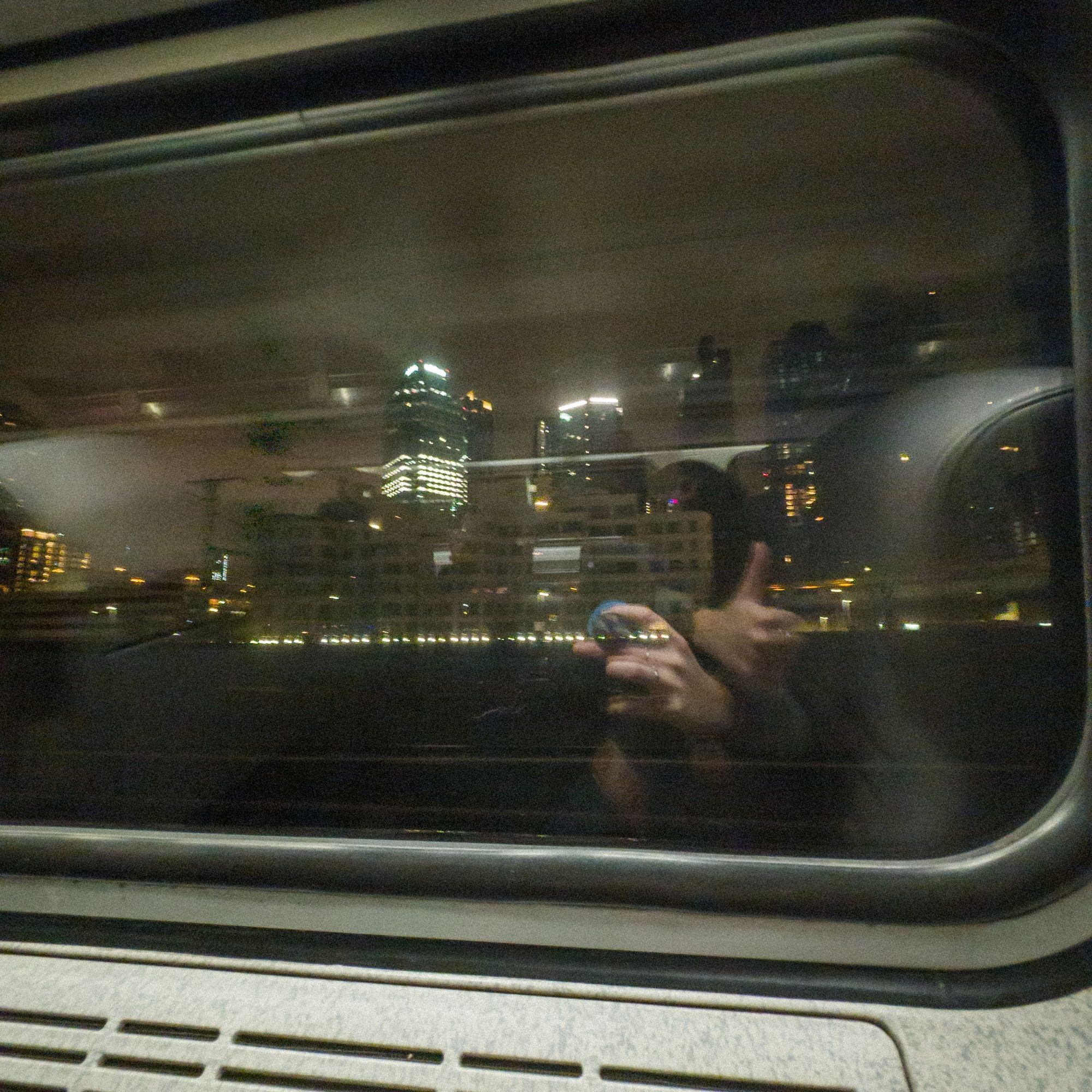 a blurry photo of the new york skyline immediately after leaving penn station viewed thru an Amtrak window at night. The hands of the photographer are reflected in the window, one hand holds a phone the other gives a thumbs up.