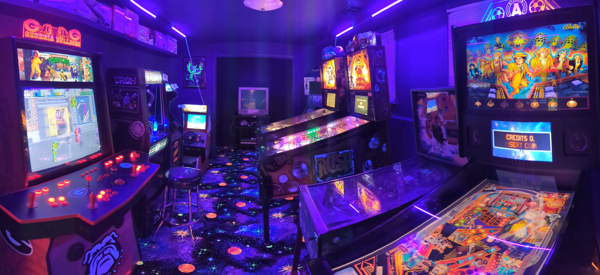 Blacklight lit, flourescent carpet, home arcade with (from left to right) a 4-player generic cab with dynamic marquee (showing TMNT), TRON Arcade1up, Star Wars Arcade1up, OutRun Arcade1up, Mandalorian Pinball (Premium), Rush Pinball (Limited Edition), Aspen Pinball (old and not working), Virtual Pinball Cabinet (showing Doctor Who)