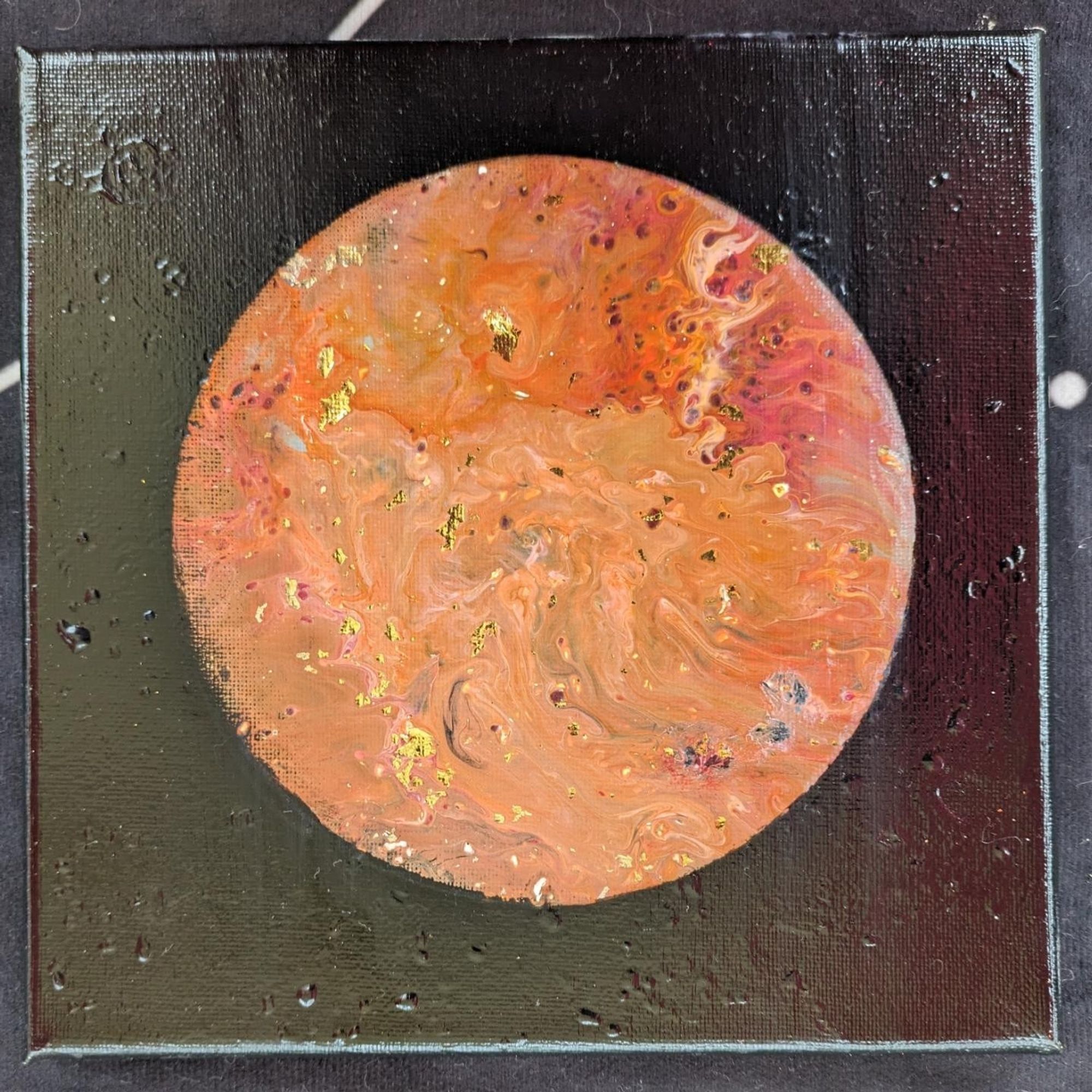 A photo of a painting on a square canvas of a planet that is orange and red, reminiscent of Mars. Gold flecks are also on the surface.